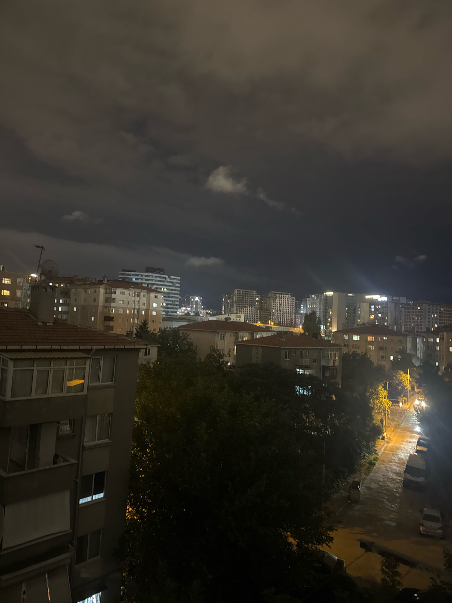 The image depicts a nighttime view of an urban landscape, likely in Gungoren, Turkey, characterized by apartment buildings and a street. The sky is dark and overcast with visible clouds. Several buildings of varying heights and architectural styles are illuminated by internal lights, indicating occupancy. In the foreground, a portion of a building is visible with windows and balconies. Beyond this, a dense canopy of dark green trees obscures parts of the mid-ground. A wet street with streetlights casting a glow on the pavement runs through the scene. Several vehicles, including what appear to be vans and cars, are parked or moving along the street. The overall impression is of a residential urban environment at night, with signs of activity despite the darkness.