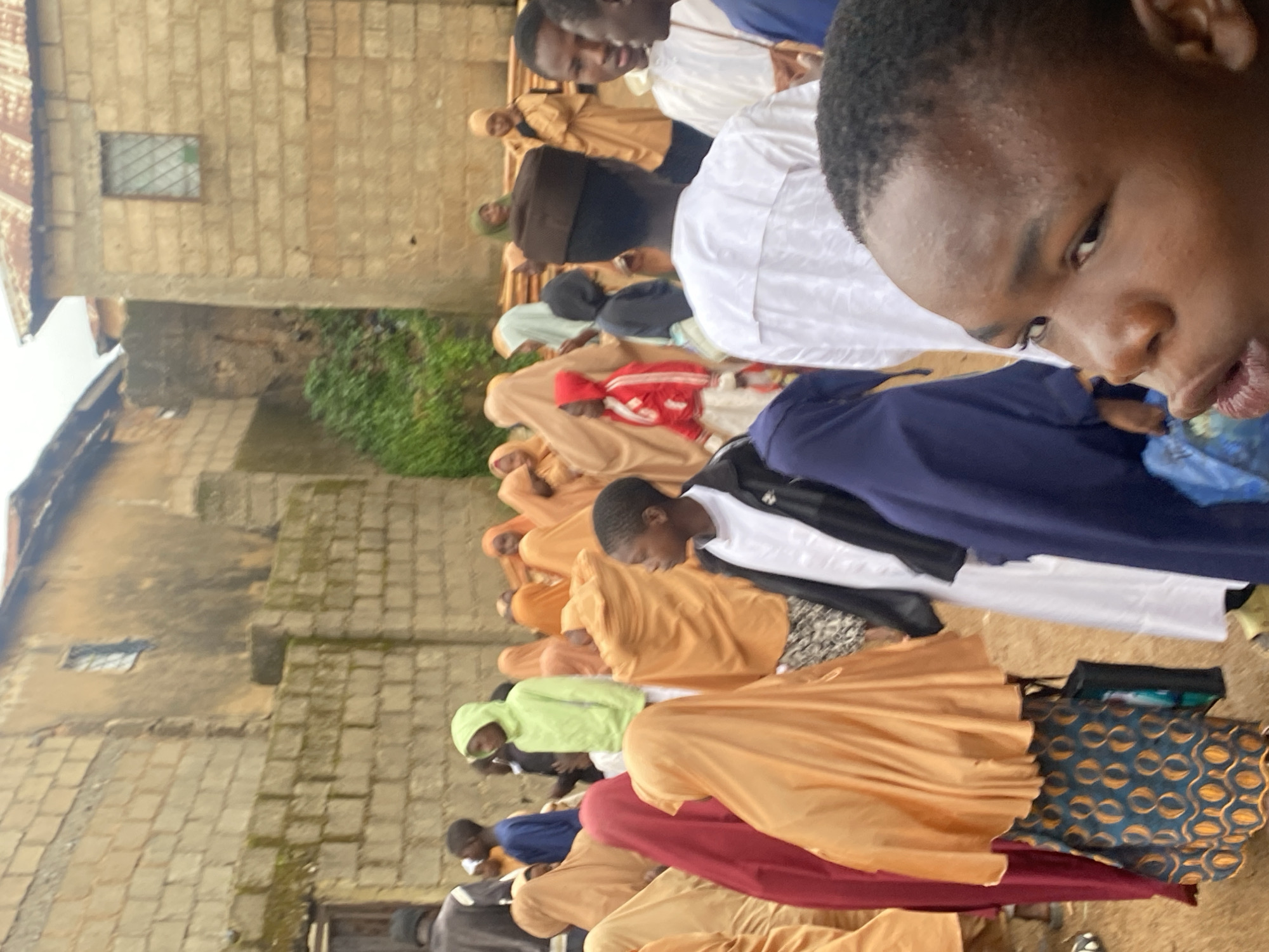 In Jos, Nigeria, on a cloudy day, a group of children and young adults are gathered outdoors in a village setting. The architecture suggests a rural or informal urban environment with buildings constructed of cinder blocks and corrugated metal roofing. Many of the individuals are wearing religious attire, with several girls and young women draped in colorful hijabs and headscarves in shades of orange, yellow, red, and blue. Some boys are wearing traditional Islamic caps and long white garments, while others are in more casual clothing like hoodies. One boy in the foreground, close to the camera, is looking off to the side with a contemplative expression. In the background, more individuals are milling about, and some appear to be engaging in conversation. The ground is dirt or unpaved, and there is a general sense of community gathering.