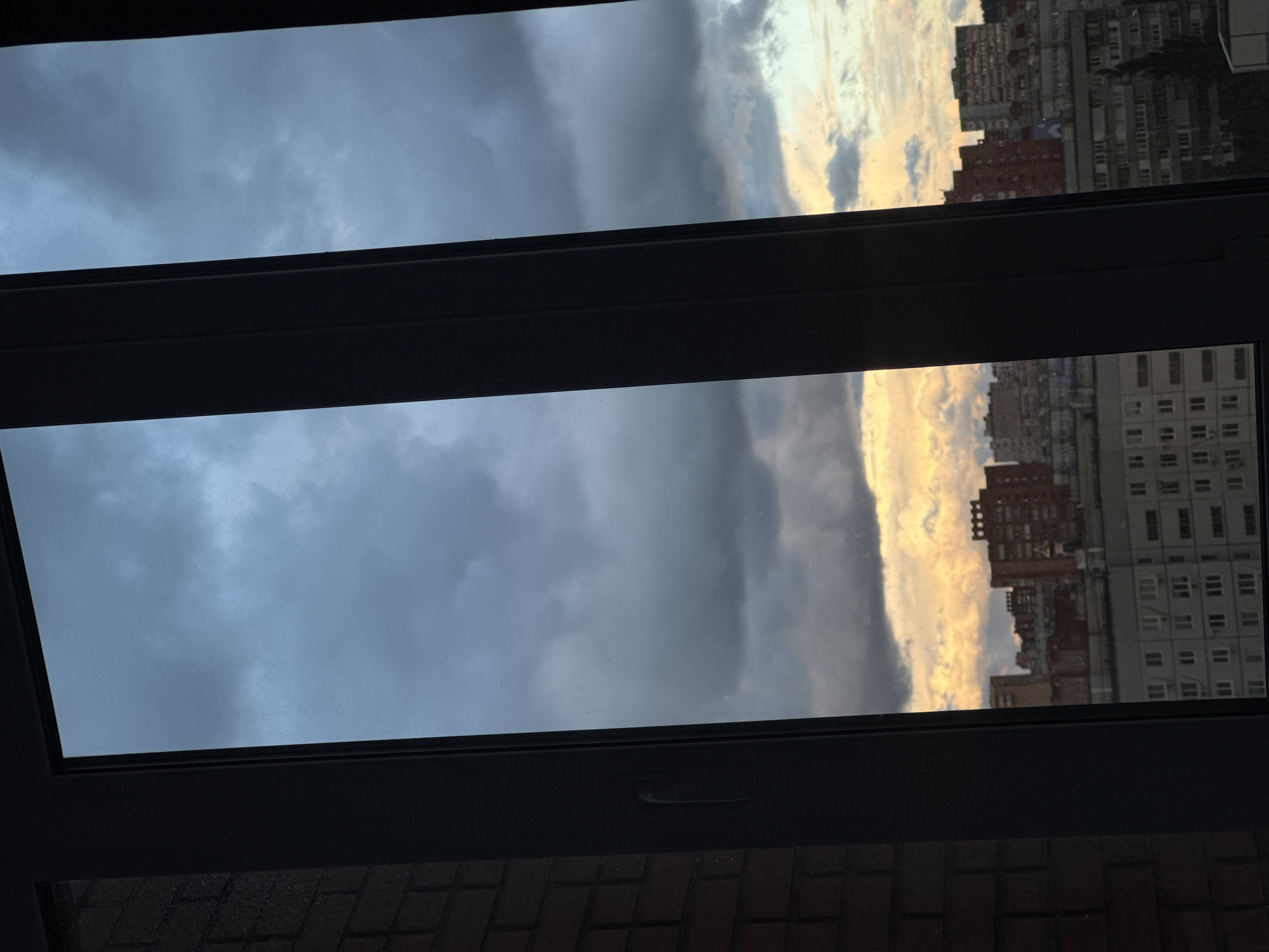 A view through an open window reveals a scene of mixed weather conditions and city structures. The left side of the window frames a sky dominated by dark, ominous clouds, indicating a potential storm or rain. Conversely, the right side shows a bright, golden sky, possibly from a setting or rising sun, contrasting with the darker clouds. Buildings are visible through the window, appearing as densely packed apartment blocks with multiple stories and windows. The window frame itself is dark and thick, providing a border to the divided sky. The window is open to a view of Unknown, Unknown. The bottom portion of the image shows a section of a brick structure, possibly a wall or part of the building where the window is located.