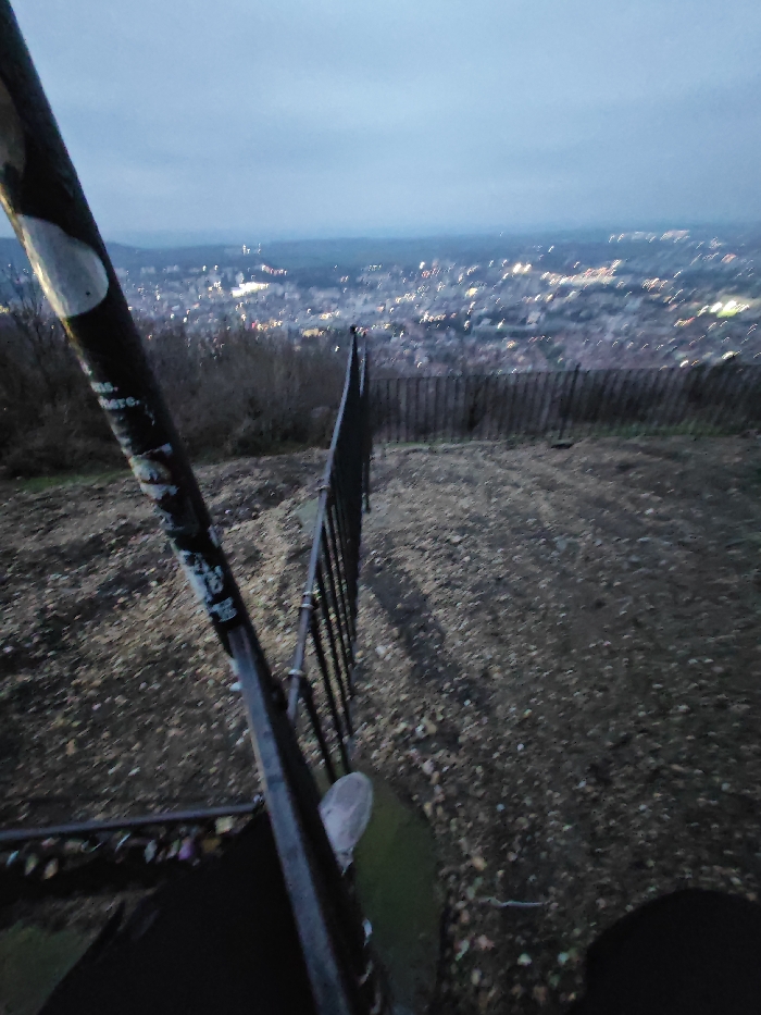 The image presents an elevated view overlooking the city of Shumen, Bulgaria, during dusk or early night conditions. In the immediate foreground on the left, a dark, angled metal post, likely part of a railing or structural support, is visible. This post features several white markings and instances of faded graffiti. The ground directly beneath and extending into the mid-ground is rough, irregular terrain covered with small fragments of debris, possibly gravel and dried leaves. A dark, segmented metal fence runs diagonally across the mid-ground, separating the foreground from a lower, slightly vegetated embankment. Beyond this fence, the urban expanse of Shumen is visible, characterized by numerous diffuse city lights illuminating buildings and infrastructure. The distant background features a faint horizon line of hills under an overcast sky, which gradually darkens towards the top of the frame. No individuals, specific actions, or interactions are discernible within the scene.