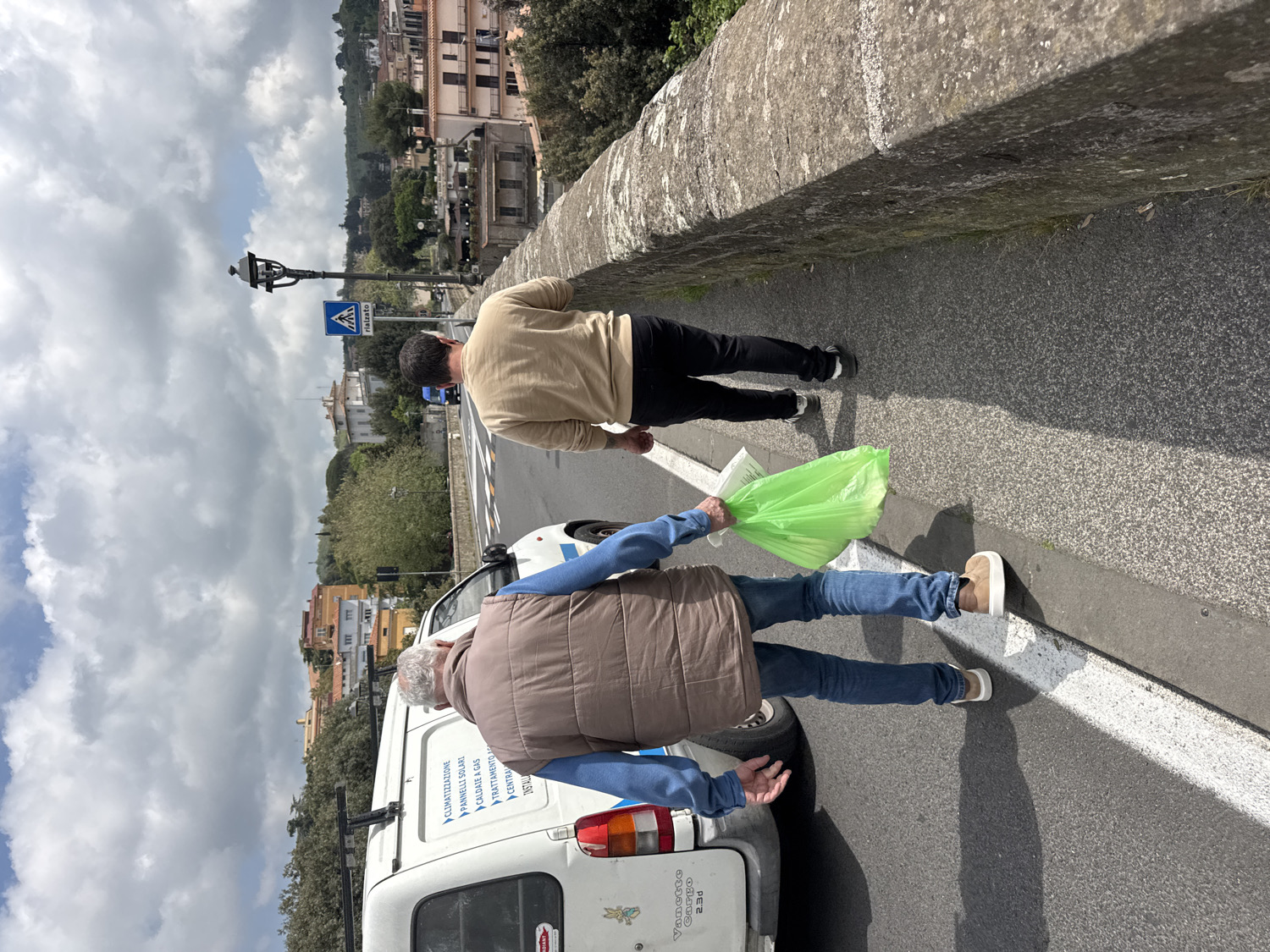 The image depicts an outdoor scene in Ariccia, Italy, featuring two men walking along what appears to be a street or elevated pathway. The sky above is partly cloudy, with patches of blue, suggesting daytime, likely late morning or early afternoon given the visible shadows.
In the foreground, a man, seen from behind, wears a blue long-sleeved shirt, a brown puffer vest, blue jeans, and light brown slip-on shoes. He holds a bright green plastic bag in his left hand, which appears to contain items. To his left, a white utility van is partially visible, parked or driving alongside the men.
Slightly ahead of him, another man, also viewed from the back, wears a light tan or beige long-sleeved shirt, dark pants, and black sneakers. Both men are walking in the same direction, away from the viewer. To the right of the men, a robust stone barrier or wall runs along the edge of the path.
In the midground and background, the scene reveals a typical Italian landscape with buildings nestled amidst green foliage and distant hills. A black lamppost stands further down the road, and a blue square road sign indicating a pedestrian crossing is visible.
Visible text on the white van includes "Vanctto Cargo 2.3d" and a list of services: "CLIMATIZZAZIONE" (Air Conditioning), "PANNELLI SOLARI" (Solar Panels), "CALDAIE A GAS" (Gas Boilers), "TRATTAMENTO" (Treatment), "CENTRA" (Central), and "INSTALLA" (Installs). A road sign in the background also shows "A rialzato," likely indicating a raised section of the road. The activity suggests a casual walk, possibly involving carrying items in the green bag. No specific emotions are discernible on the subjects.