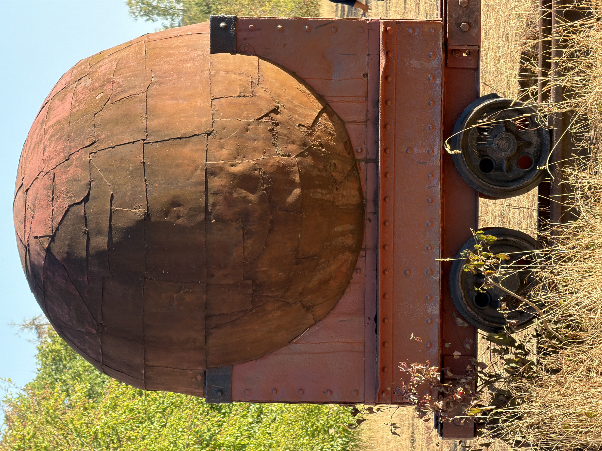 The image depicts a large, weathered, spherical sculpture, appearing to be made of rusted metal panels, mounted on a red, industrial-style trolley. The trolley has two visible wheels, and the entire structure is situated outdoors in a dry, grassy field. Behind the sculpture, there are trees with green foliage and a clear blue sky. A person's arm is visible on the far right edge of the image, suggesting their presence near the sculpture. The sculpture is the primary focal point, with its rough texture and segmented surface clearly visible. The scene suggests an outdoor art installation or a piece of industrial heritage. The sculpture is located in Schifflange, Luxembourg, specifically at the Fond-de-Gras, a historical mining site that has been transformed into a tourist attraction and museum.