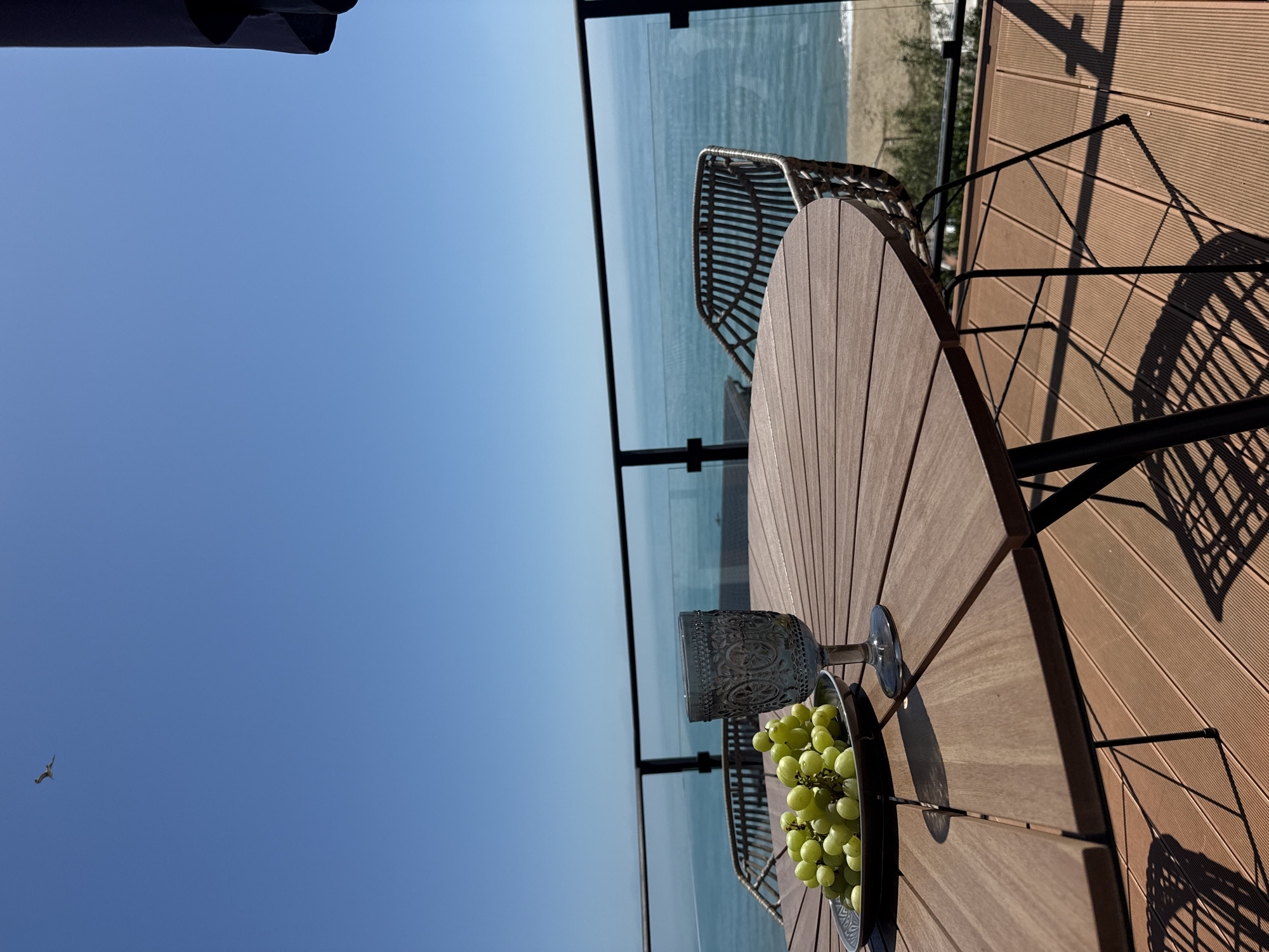 An outdoor scene captured on a sunny day, likely in Varna, Bulgaria. A round wooden table stands on a brown wooden deck with metal chairs, overlooking the Black Sea through a glass railing. On the table is a bowl filled with green grapes and a decorative glass. The blue sky is mostly clear with one bird flying in the lower left corner. The scene is bathed in bright sunlight, casting shadows of the chairs on the deck.