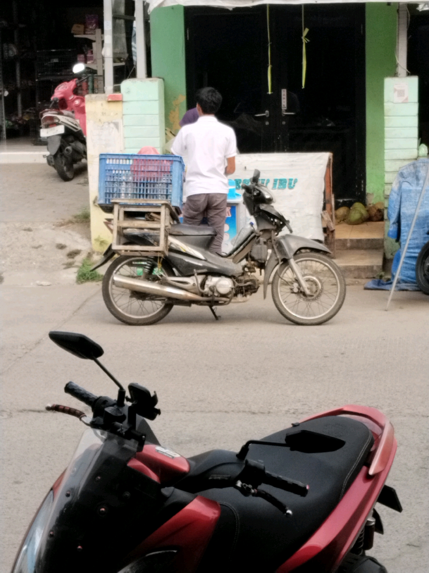 In Serang, Indonesia, a man in a white shirt and grey trousers stands next to a dark motorcycle parked on an asphalt surface. The motorcycle has a wooden crate and a blue plastic basket secured to its rear. In the background, a pink motorcycle is visible to the left, and a building with green and light blue accents stands to the right. The scene appears to be an outdoor market or roadside stall.