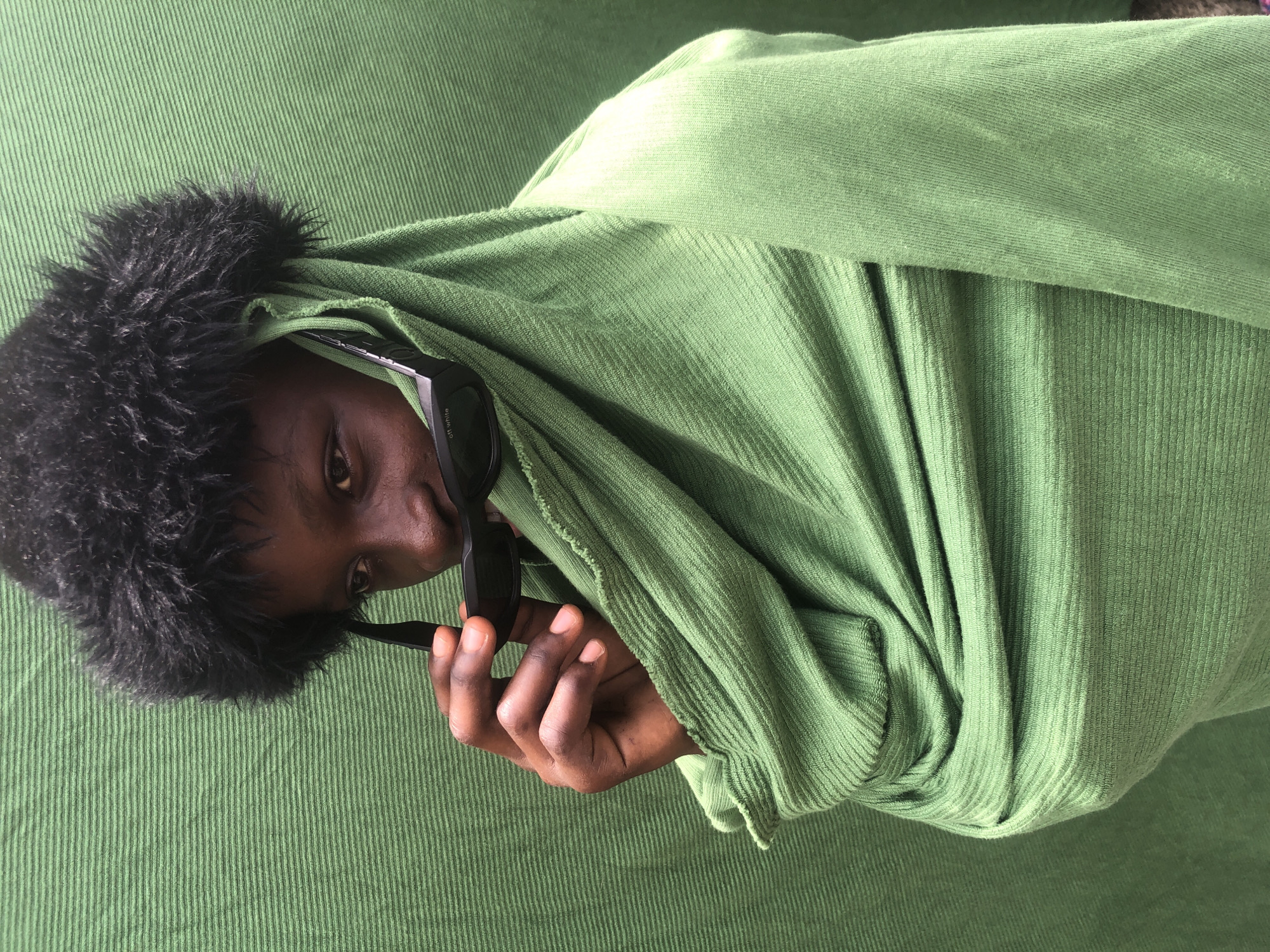 A close-up shot features a young person wearing a fuzzy black hat and a draped green garment. They are holding a pair of black sunglasses up to their face, obscuring their mouth and nose. The background is a textured green surface. The image appears to be taken outdoors in a dimly lit setting, possibly in Ado, Nigeria, based on the user's prompt. The lighting is soft, creating subtle shadows and highlighting the textures of the clothing and background. The person's eyes are visible, looking slightly to the side. The overall mood is mysterious and artistic.