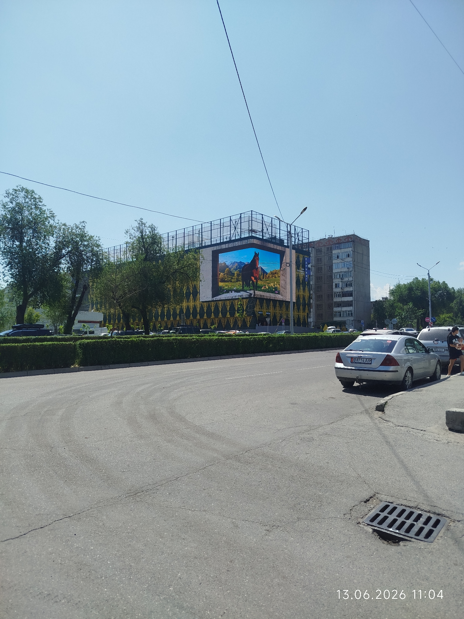 The image captures a daytime scene on a city street, likely in Bishkek, Kyrgyzstan, as indicated by the timestamp. A large LED billboard displaying a horse against a mountainous backdrop is prominently featured. Adjacent to the billboard is a multi-story apartment building. The foreground shows a paved road with a slight curve, a storm drain, and a sidewalk. Several cars are parked or in motion on the street, including a silver sedan with license plate "B8714 AR". Two individuals, a woman holding a child, are standing on the sidewalk near the parked cars. The environment appears to be a developed urban area with trees lining the street and a neatly trimmed hedge separating the road from the billboard and buildings. The sky is clear and blue.