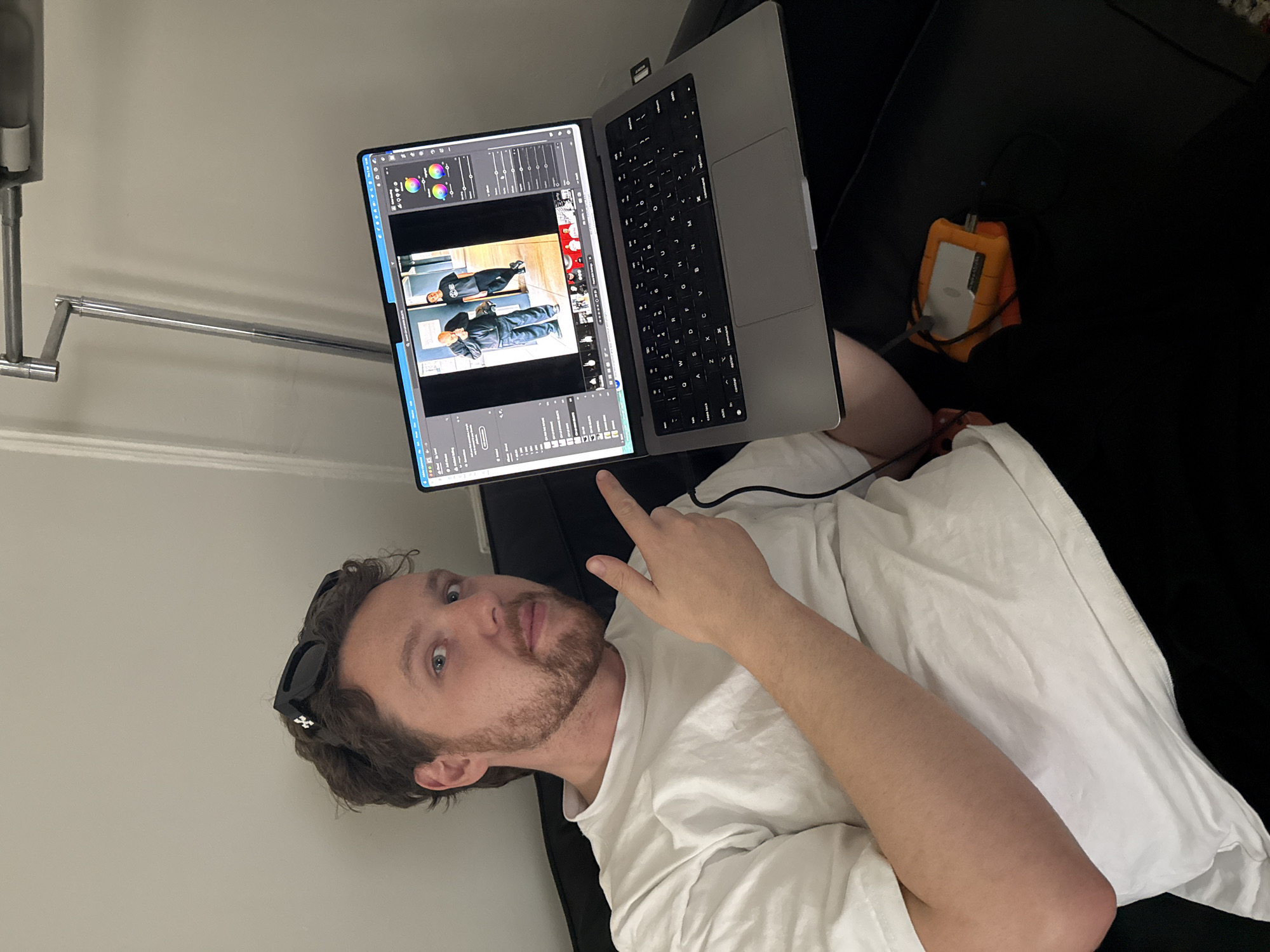 A man with stubble and sunglasses atop his head reclines on a black couch, pointing up at a silver laptop which appears to be running photo editing software. An orange external hard drive is plugged into the laptop. The man is wearing a white t-shirt and is partially obscured by a white pillow. A silver articulated lamp arm is visible in the background. The environment appears to be an interior space. The location and city cannot be determined from this image.