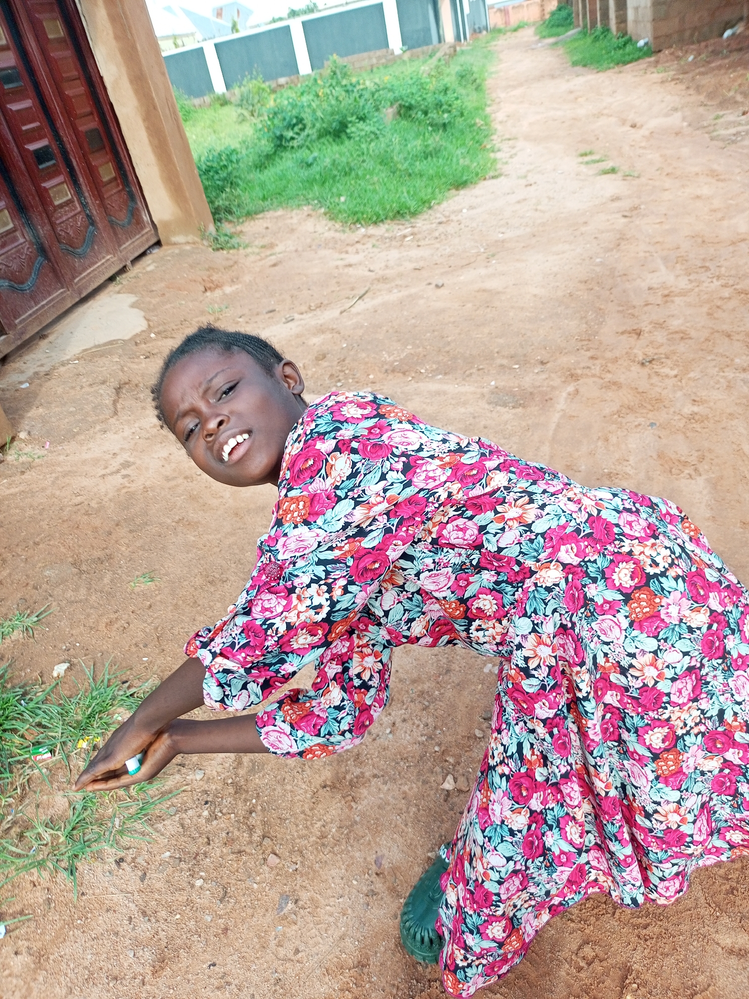 A young Black girl in a floral dress and green shoes is shown from the waist up, bending down and looking at the camera with a playful expression. Her left hand is held near the ground, and it appears she is touching a small, white object, possibly a piece of candy or a pen. The background shows a dusty path, some green grass, and a brown wall with a large, red double door to the left. In the distance, there are some buildings and more greenery. The lighting suggests it is daytime. The location is identified as Tempure, Nigeria.