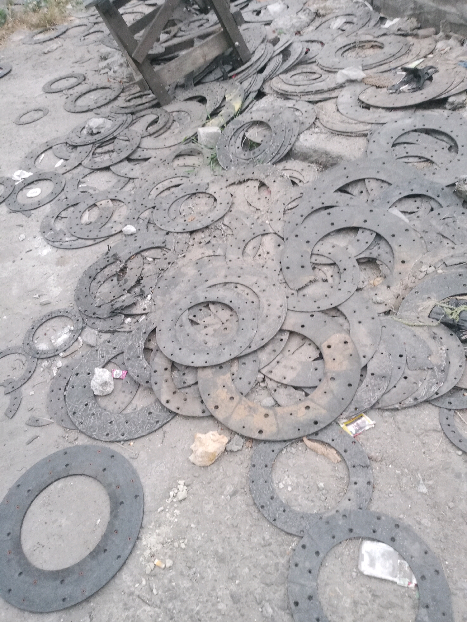 The image depicts a scene in Balaraja, Indonesia, featuring a large collection of worn, circular, flat objects, possibly clutch plates, scattered across a grey, dusty ground. The objects are dark grey and exhibit numerous small holes punched around their circumference and some in the center. They are piled haphazardly, with some overlapping and others lying flat. The ground appears to be a mixture of dirt, sand, and small stones. In the background, a wooden structure, possibly part of a makeshift stall or scaffolding, is visible, along with some dry vegetation. There are no people present in the image. The overall impression is one of a junkyard or a storage area for discarded industrial or automotive parts.