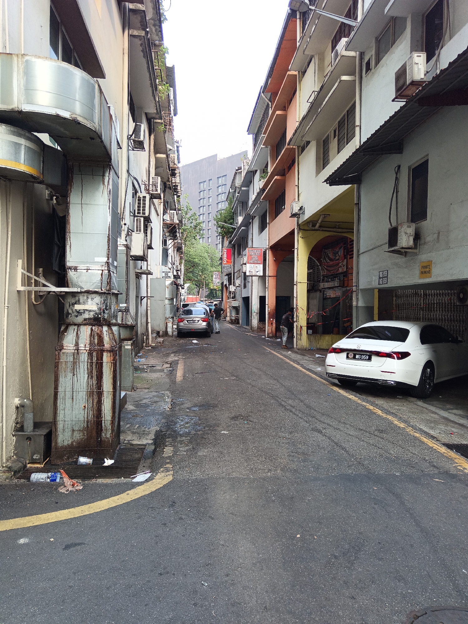 The image depicts a narrow alleyway in Kuala Lumpur, Malaysia, characterized by a row of multi-story buildings on either side. The buildings exhibit a mix of architectural styles and varying states of maintenance, with some featuring faded paint and visible signs of wear. Large, rusted metal ducts extend from the side of a building on the left, dominating the foreground. The alleyway is paved with asphalt and marked with a yellow line. Litter, including plastic bottles and wrappers, is scattered on the ground.
Several vehicles are present in the alley. A white sedan is parked in a covered area on the right, its license plate clearly visible. Further down the alley, a silver sedan is parked, with two individuals standing near it. The individuals appear to be engaged in a conversation or some form of interaction. The overall environment suggests an urban setting, likely a commercial or residential area with limited public space. The sky is overcast, and the lighting suggests it is daytime.