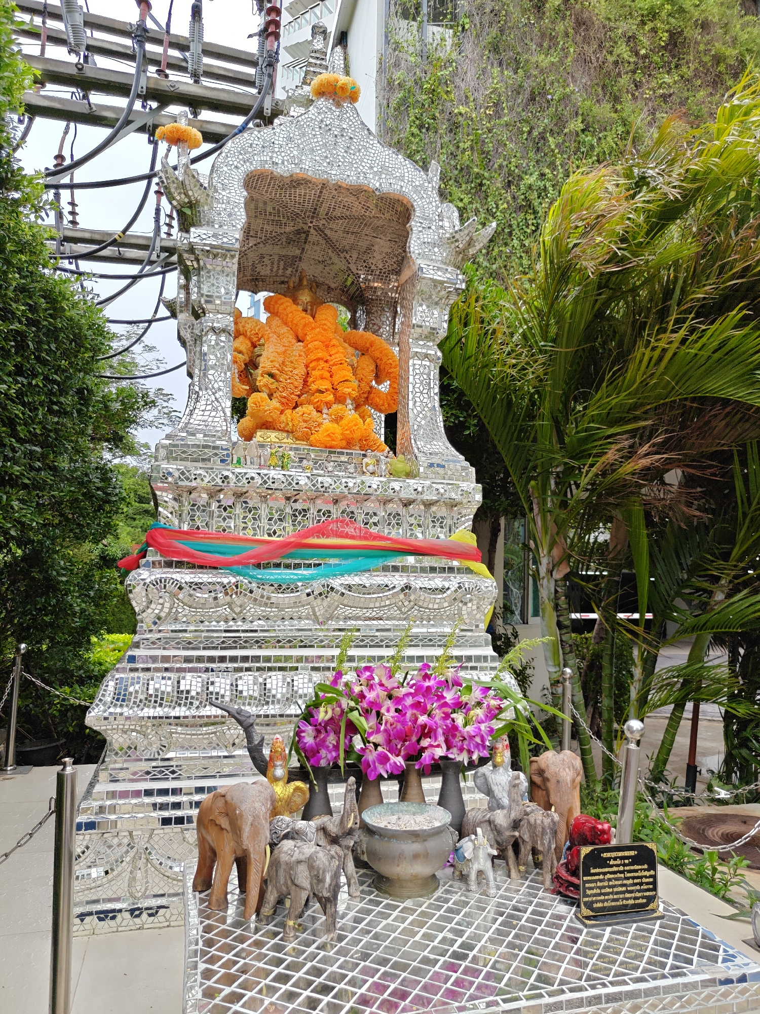This is a full-shot, eye-level, outdoor, daytime photograph of a shrine in Pattaya City, Thailand. The shrine is elaborately decorated with mirrored tiles and adorned with offerings. The shrine itself is a multi-tiered structure with intricate carvings and embellishments. At the top, a golden statue is visible, draped with vibrant orange flower garlands. The shrine is decorated with colorful fabric ribbons in red, yellow, and green. In front of the shrine, on a mirrored surface, are several wooden animal figurines, including elephants and a rooster, as well as a vase of pink orchids and an incense burner. A dark plaque with Thai text is also present. The shrine is situated amidst lush greenery, with palm trees and other tropical plants visible. Electrical power lines and poles are present in the background, indicating an urban or semi-urban setting. The overall impression is of a sacred space decorated for veneration.