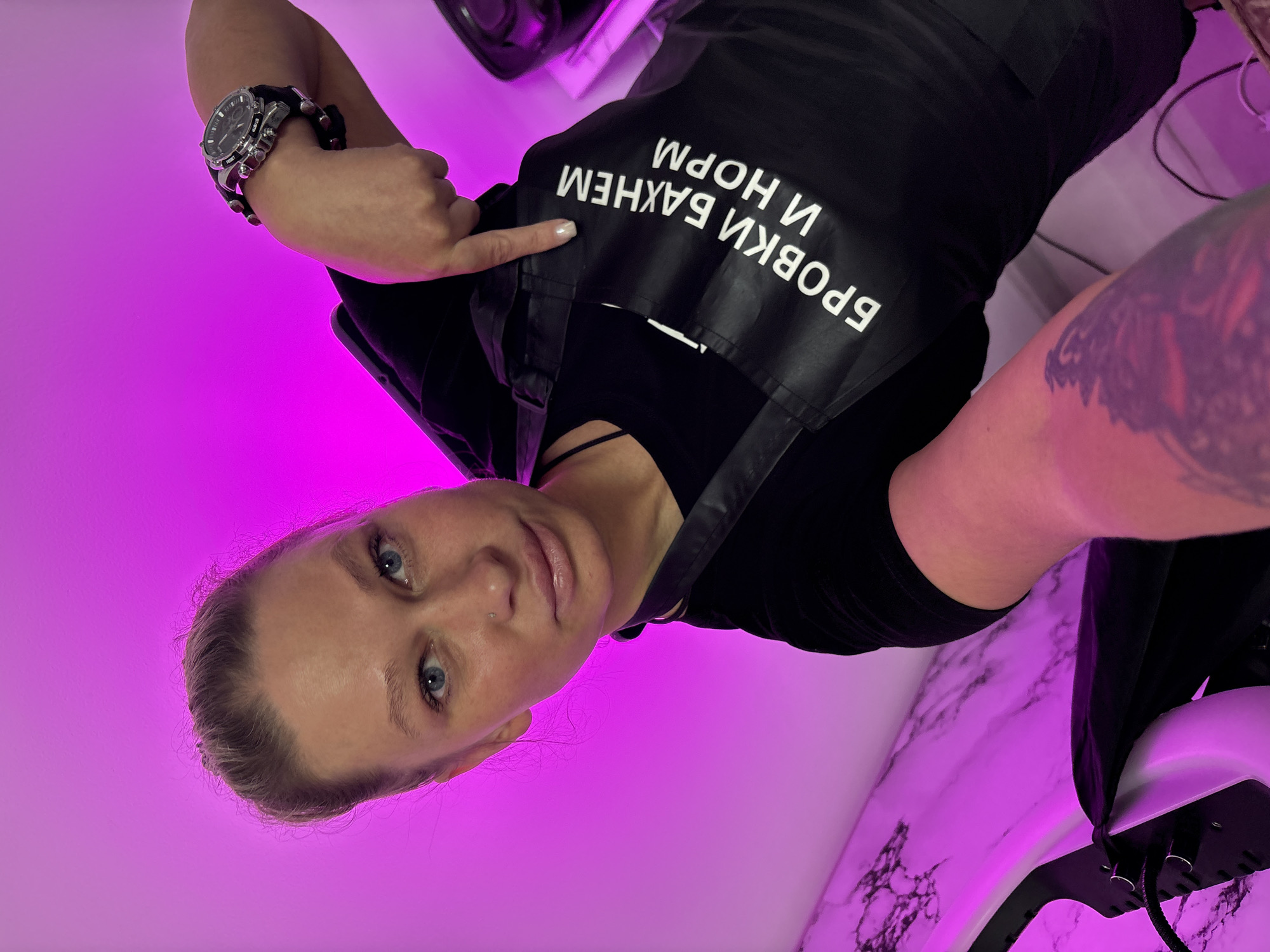 A woman is captured in a close-up, selfie-style portrait, with the image oriented vertically as if taken from below and looking up, then rotated. She has fair skin, light-colored hair pulled back from her face, and blue eyes. A small stud nose piercing is visible on her left nostril. She wears a black top beneath a black apron or vest that features white Cyrillic text.
Her left hand is raised, with the index finger pointing towards the text on her garment. On her left wrist, she sports a prominent multi-dial analog watch with a dark strap and silver-toned casing. A dark, intricate tattoo covers her right upper arm and shoulder.
The white Cyrillic text on her black apron, when properly oriented, reads "БРОВКИ ВАЖНЕИ И НОРМ," which translates to "BROWS ARE IMPORTANT AND NORMAL" or "BROWS ARE MORE IMPORTANT AND NORMAL." The background is illuminated by a strong magenta-purple light, indicating an artificial lighting setup. In the lower right of the frame, a white surface with black marbled patterns is visible, along with a portion of a white, dome-shaped device, possibly a beauty appliance. The woman looks directly at the camera with a neutral or slightly smiling expression. The setting appears to be an indoor environment, likely in Perm, Russia.