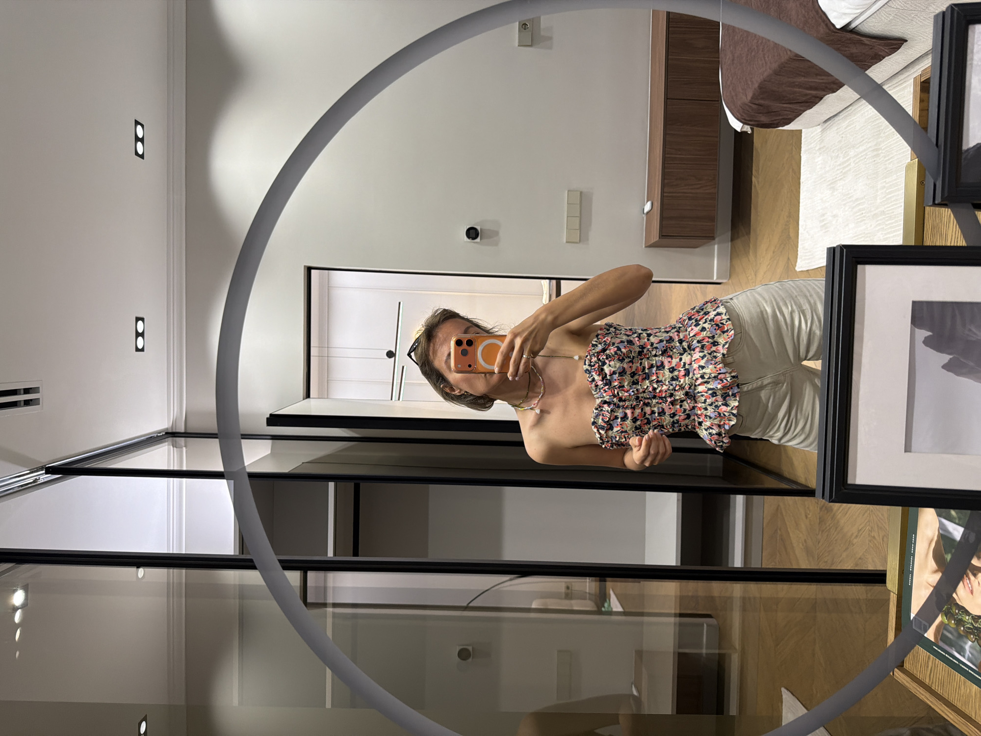 A woman stands in a room, taking a mirror selfie. She is wearing a floral print strapless top and light-colored shorts. In her right hand, she holds an orange smartphone, angled to capture her reflection. She is wearing a necklace made of small beads and a single pearl. The room appears to be a bedroom or dressing area, with a large mirror reflecting the scene. Behind the woman, a closet with glass doors is visible, as well as a bed with a brown comforter. The walls are painted a neutral color, and recessed lighting is installed in the ceiling. The floor is made of wood. There are framed pictures on a table in the foreground.