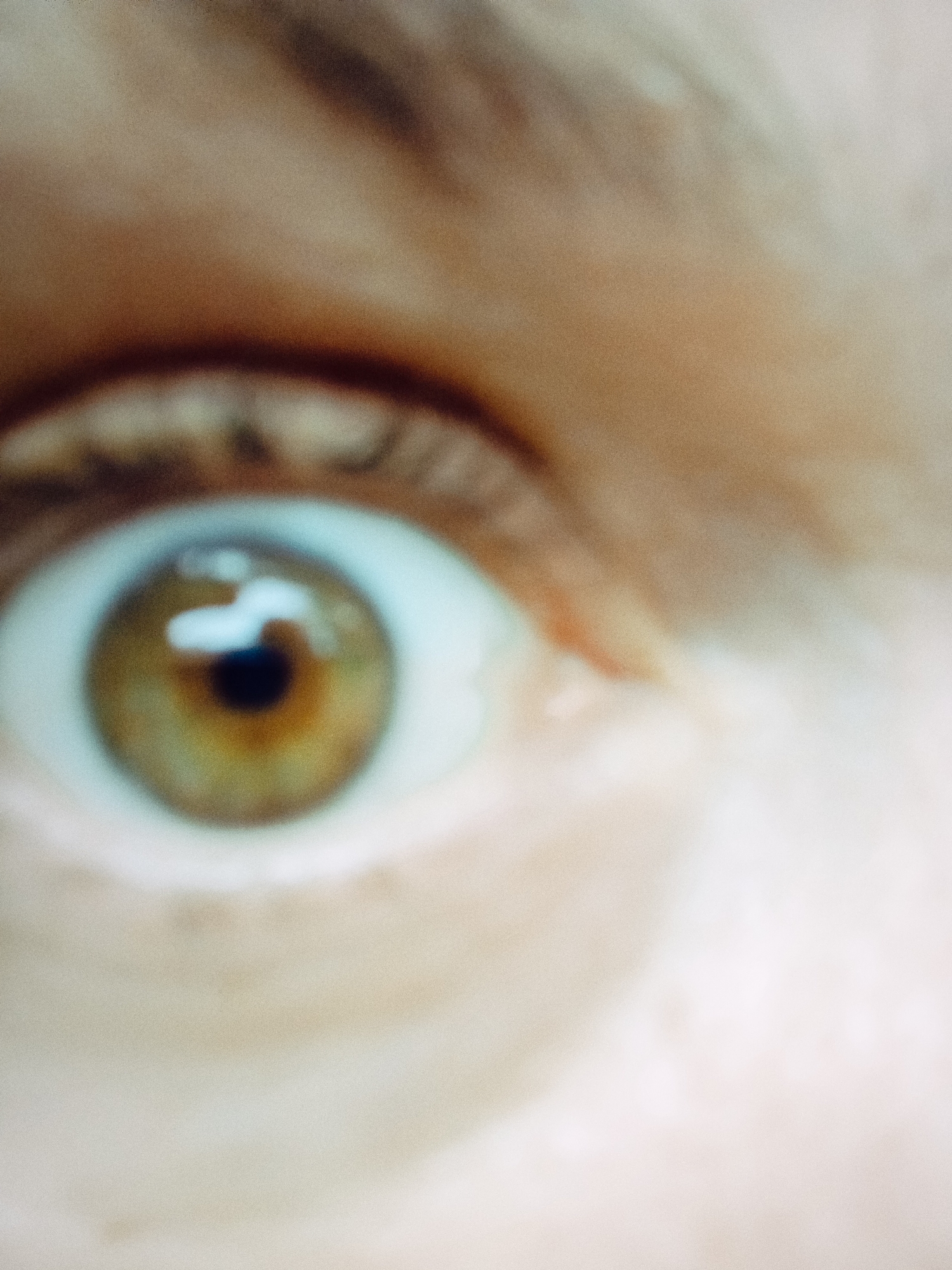 A close-up, blurred image of a human eye with a light brown iris. The surrounding skin and eyebrow are also visible, though out of focus. The location and city are not discernible from this image.