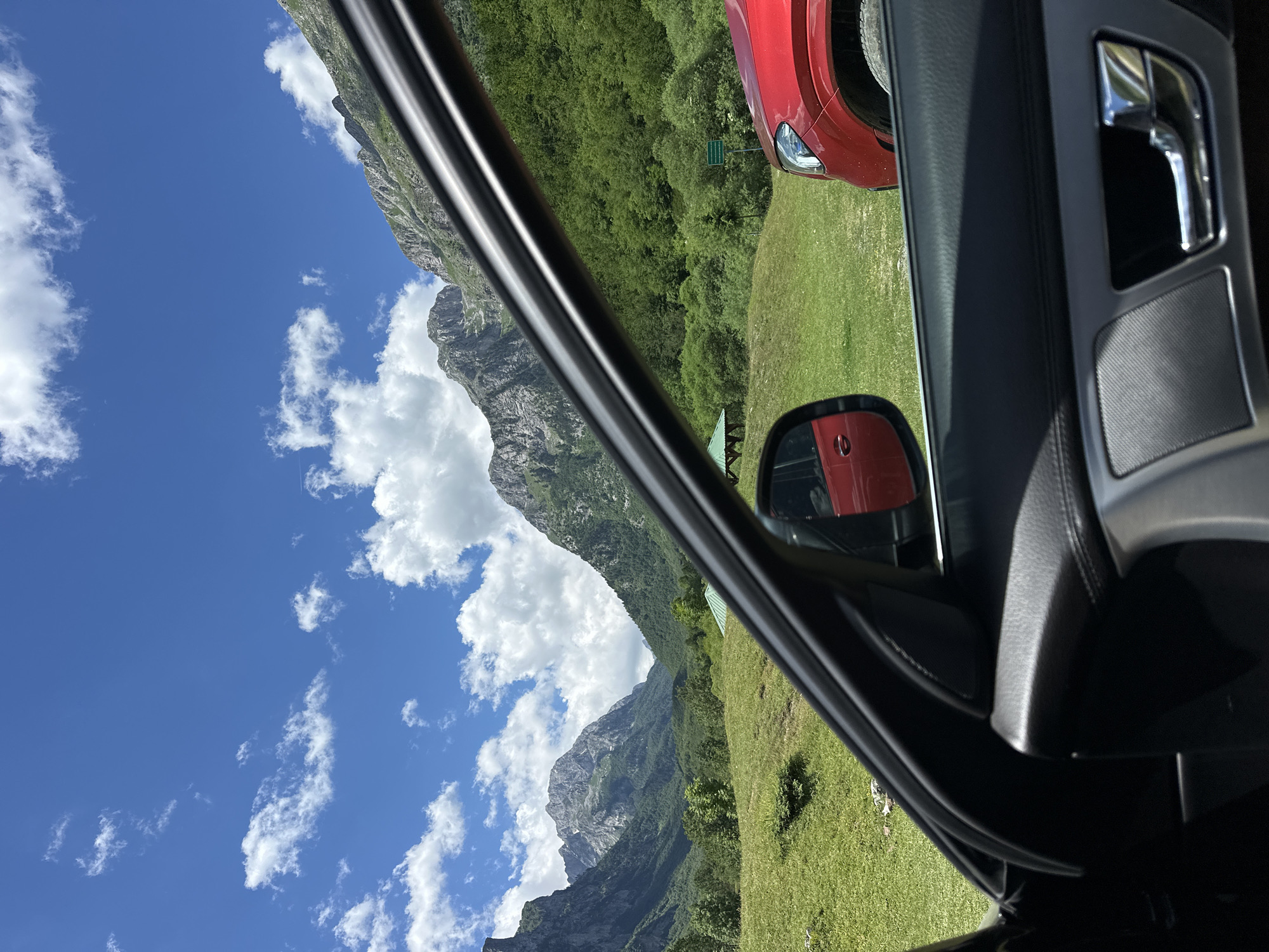 The image captures a view from inside a car, looking out at a mountainous landscape under a bright blue sky with scattered white clouds. In the foreground, the interior of the car is visible, including a portion of the door panel with a silver door handle and a speaker grille. A side-view mirror reflects a red car.
The exterior scene features rolling green hills, densely forested slopes, and rugged, rocky mountain peaks in the distance. A small, dark-colored gazebo or shelter with a green roof is situated on the grassy slope. Another red vehicle, possibly an SUV, is parked on the right side of the frame. The overall environment suggests a rural or natural setting, likely in a mountainous region like the area around Gusinje, Montenegro, known for its dramatic scenery. There are no people visible in the image.