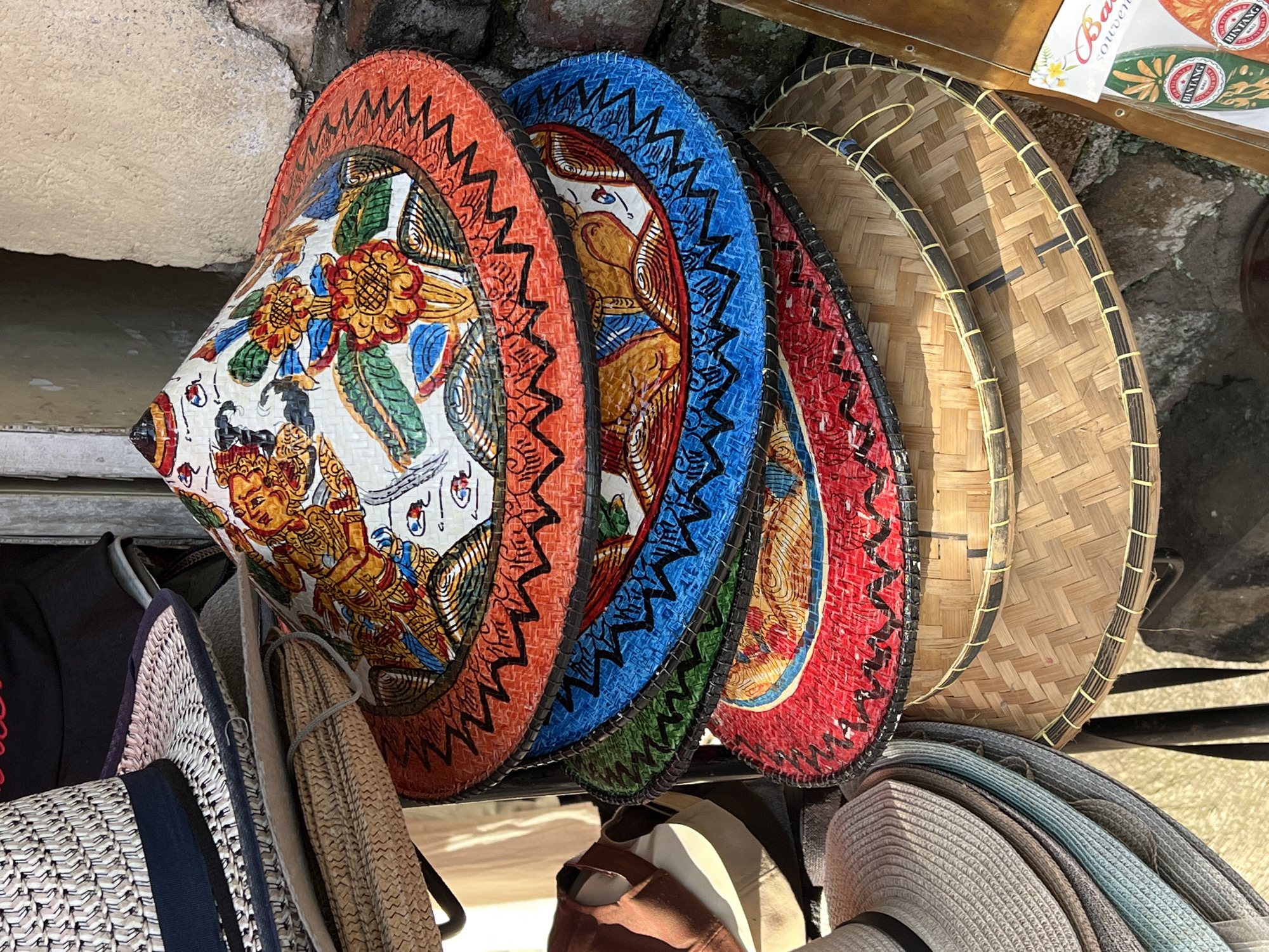 The image captures a display of colorful, conical Asian hats in Ubud District, Indonesia. The hats are densely arranged on what appears to be a rack or display stand. The front-most hat is predominantly white with intricate hand-painted designs in shades of orange, yellow, blue, and green, featuring a depiction of a figure in golden attire. Subsequent hats display solid colors of orange, blue, green, and red respectively, with black zigzag borders. Behind these are three woven straw hats. A collection of other hats is beneath the conical hats. The background shows a stone-like wall.