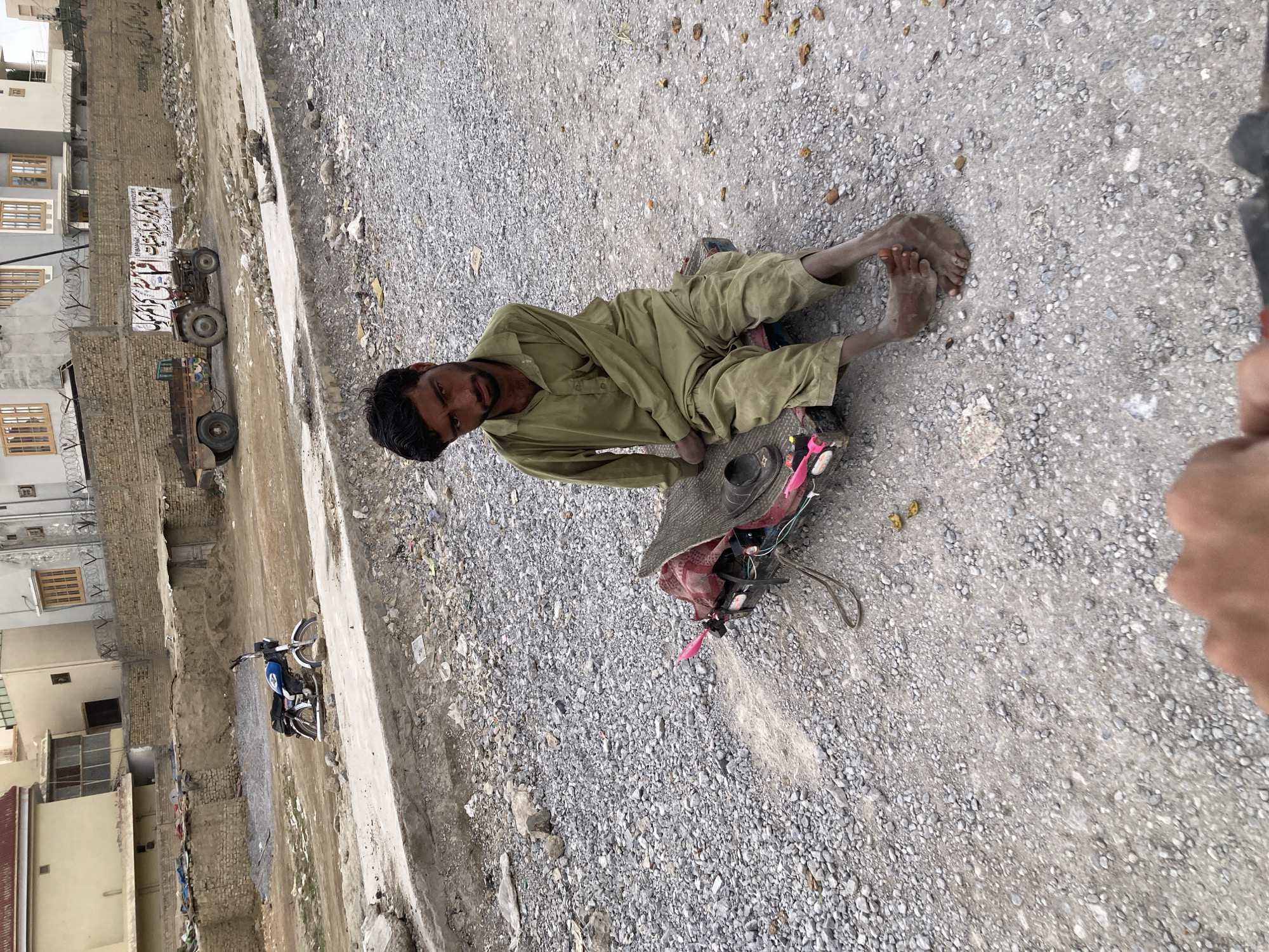 This media file presents a rotated, eye-level view of a street scene in what appears to be Quetta City Tehsil, Pakistan, as indicated by the location context and visible script.
The primary subject is a man of South Asian descent, dressed in a light olive-green shalwar kameez, sitting on the gravel and dirt ground. He is facing slightly towards the camera, with a neutral, possibly weary expression. His feet are bare and appear dirty. Next to him lies a dark, wide-brimmed hat, beneath which some pink fabric or objects are visible. A small, dark bowl or cup is also on the ground near his right hand. The immediate ground is covered in small gray gravel and dirt, with some scattered debris.
In the background, to the left of the man (as the image is rotated), a dirt road or unpaved area extends, bordered by a concrete curb or wall. Beyond this, a row of brick and concrete buildings is visible. Many windows are visible, and some buildings feature barbed wire along their tops for security. There appears to be a construction site or an area with exposed foundations and rubble between the buildings and the dirt path.
Several vehicles are present in the background. A small tractor with a white trailer or cargo bed is prominent, featuring clear Urdu script. The text reads "بسم اللہ الرحمن الرحیم" (Bismillah-ir-Rahman-ir-Rahim - In the name of Allah, the Most Gracious, the Most Merciful), followed by "پٹرولیم" (Petroleum) and "سروس" (Service), suggesting it might be used for transporting fuel or related services. Another, smaller truck-like vehicle is partially visible behind the tractor. Further down the path, a blue and white motorcycle is parked.
The scene takes place during daylight hours, under what appears to be an overcast sky, as shadows are soft and diffused. The general atmosphere suggests a non-affluent, possibly working-class or semi-rural urban environment. The man's posture and items suggest he might be resting or engaged in begging.