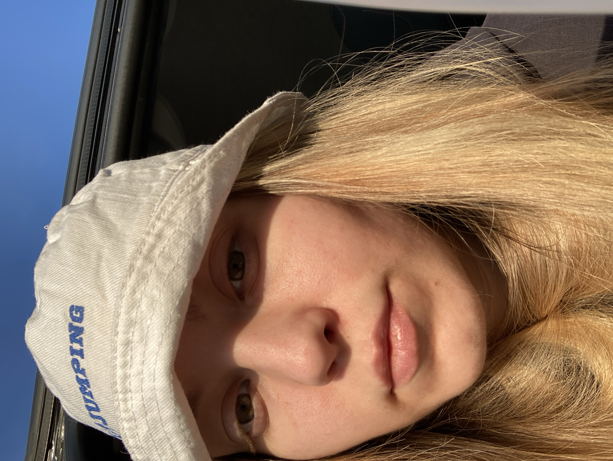 A close-up portrait of a fair-skinned person with long blonde hair is wearing a white bucket hat with the word "JUMPING" printed vertically in blue on the left side. The individual is positioned indoors near a window or opening with a view of the sky. The sky appears blue. The lighting is strong, creating shadows across their face. The person is facing the camera with a neutral expression. The background shows a partial view of a dark, possibly black, interior frame.