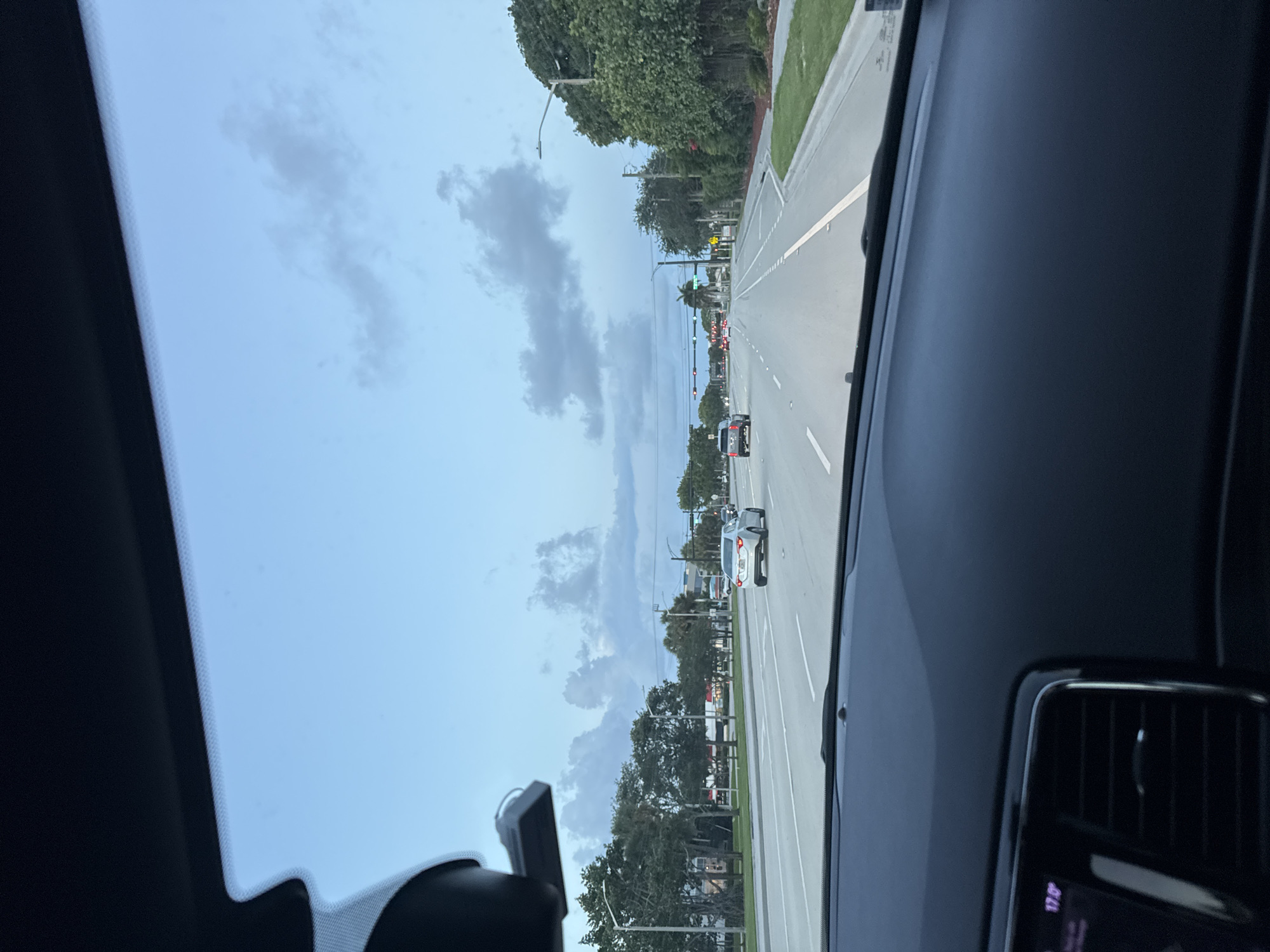 The image shows a view from inside a car, looking out onto a road in Boca Raton, Florida. The sky is overcast with scattered clouds. The road has multiple lanes with a center median and is lined with trees and grass. Traffic is visible in the distance. A rearview mirror and dashboard elements are visible inside the car.