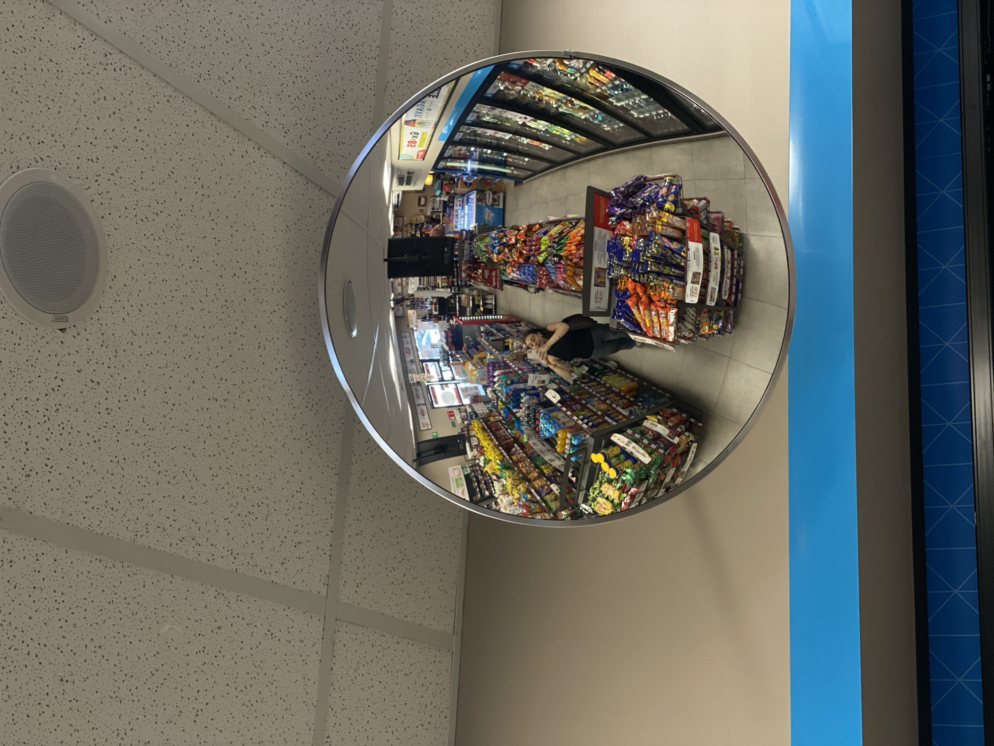 The scene captured in the image is the reflection in a convex mirror inside a convenience store, presumably for security purposes. The reflection shows a view of the store's interior, with aisles of snack foods and beverages. A person is standing in one of the aisles, possibly looking at or reaching for items. The mirror is attached to a wall that transitions to a blue panel, possibly part of a display case or refrigeration unit. Above the mirror, there is a textured white ceiling with a circular speaker. The location is a convenience store in the city of Unknown, Unknown.