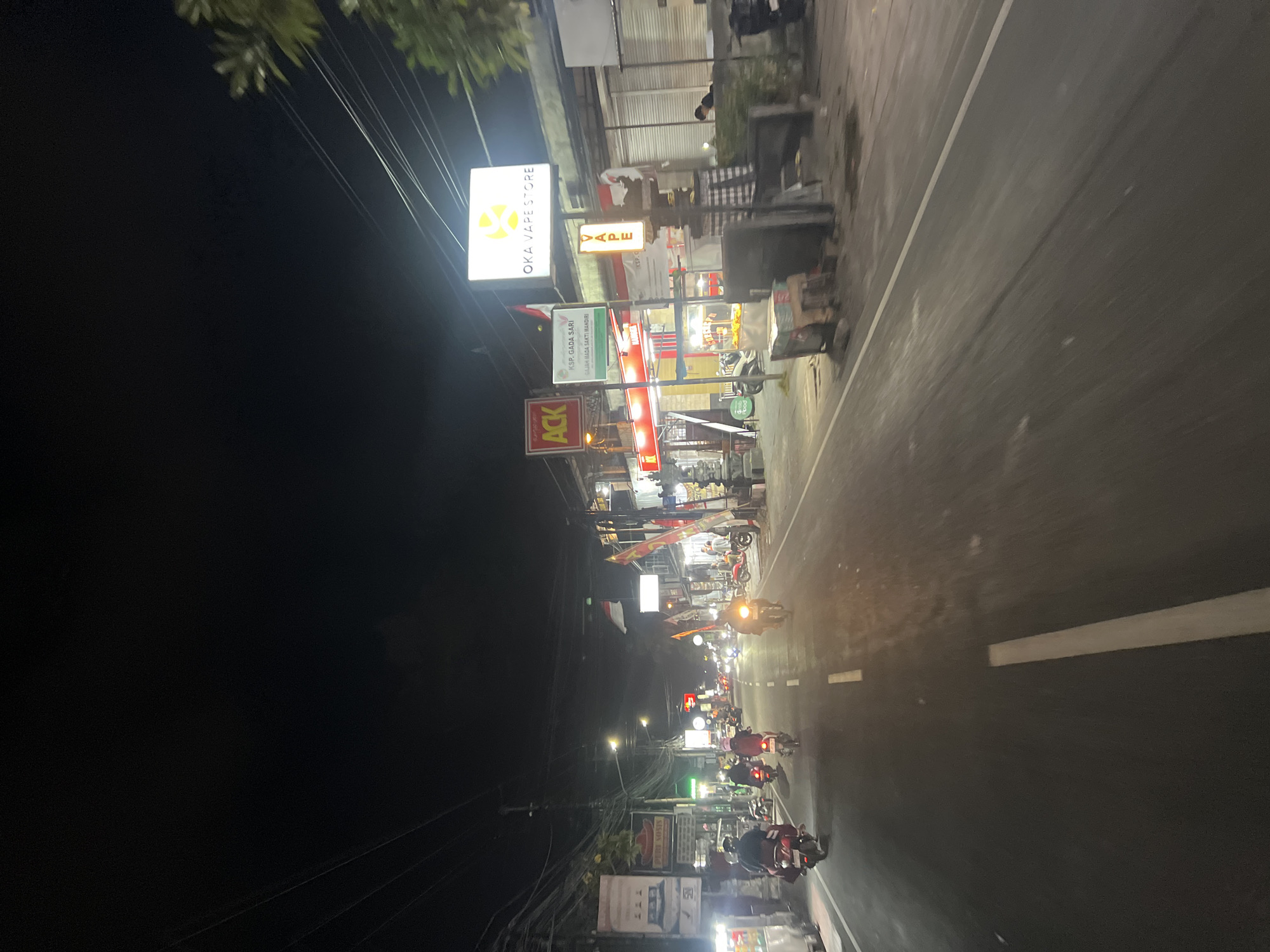 The image depicts a street scene at night in Denpasar, Indonesia, likely along a commercial strip. The perspective is from a moving vehicle, possibly a motorcycle, looking forward down the road.
On the right side of the street, a brightly lit sign reads "OKA VAPE STORE" with a stylized logo. Below this, a smaller vertical sign displays the word "VAPE." Further down the street, other illuminated signs are visible, including one with the text "ACK." Some buildings appear to be shops with their interiors lit. There are also decorative elements, such as a traditional Balinese checkered cloth hanging near the vape store.
On the left side of the road, several motorcycles are visible, some with riders. One rider is wearing a red jacket. The road itself has white lane markings. The sky above is dark and appears to be the night sky, with some power lines crisscrossing the upper portion of the frame.
The overall impression is a bustling, well-lit street at night, characteristic of a commercial area in an Indonesian city.