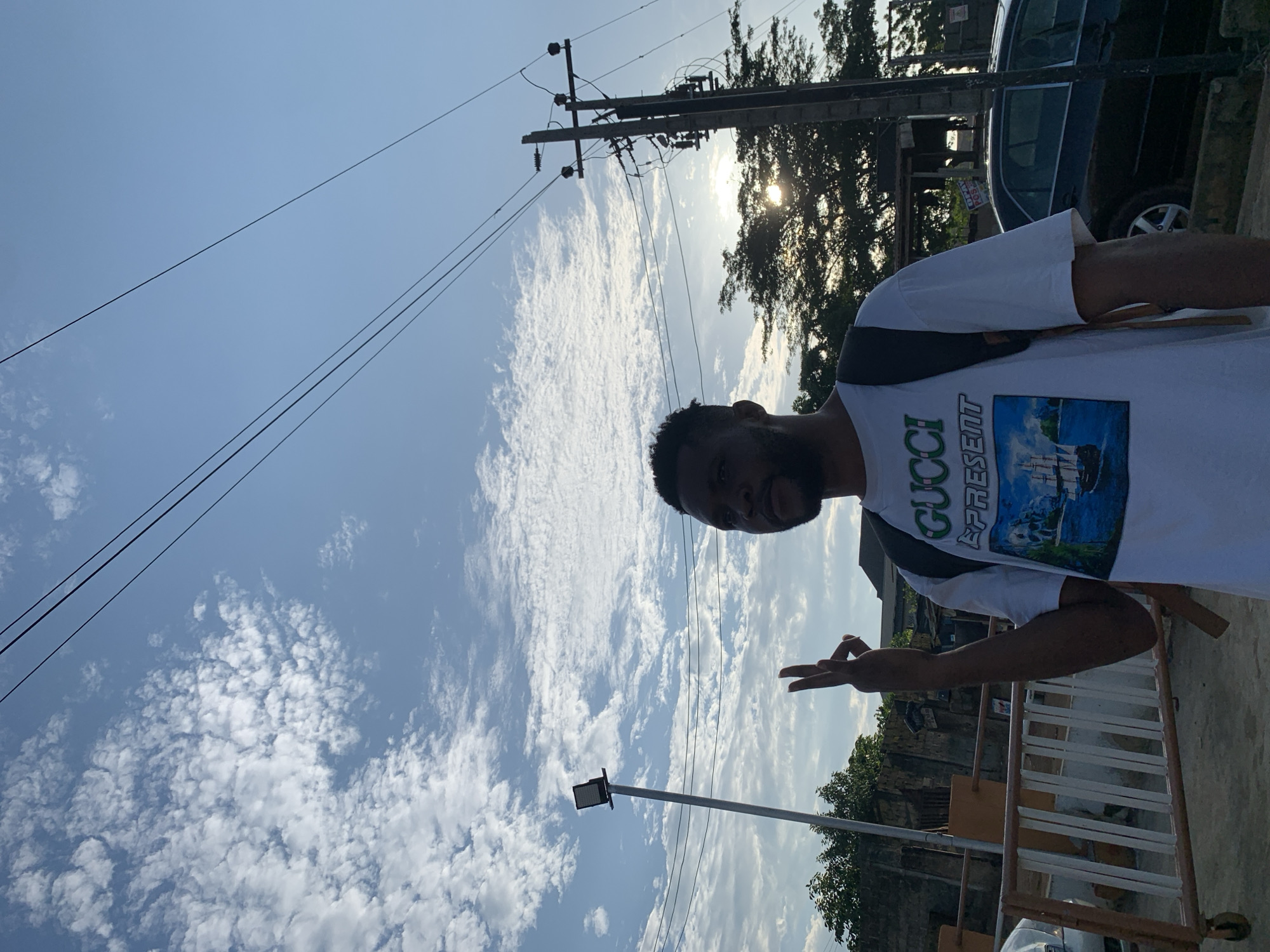 A man stands in Ikorodu, Nigeria. He wears a white T-shirt with "Gucci Represent" printed on it. Above him is a blue sky with scattered clouds and power lines. A utility pole is visible in the background, along with trees and a car. The man is making a hand gesture. A streetlight pole and metal barriers are also visible.