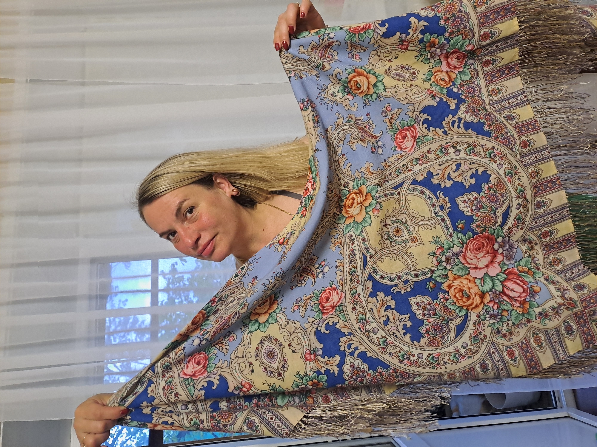 A blonde-haired woman is holding up a large, colorful shawl with a floral pattern and fringed edges. The shawl has a predominantly blue and yellow color scheme with intricate floral designs. The woman is looking towards the camera with a slight smile. The background appears to be a room with a window. Judging from the window light it is daytime.