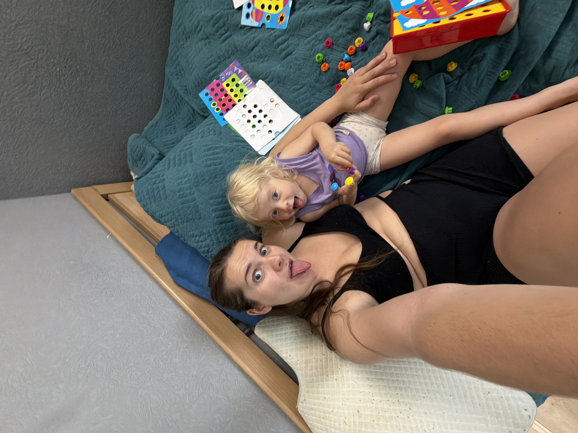 A selfie shows a woman and a child lying on a bed, likely indoors. The woman, in a black top and shorts, has brown hair and is sticking her tongue out, making a silly face. The child, dressed in a purple shirt and light-colored shorts, is also making a face while holding colorful toys. The bed is covered with a blue quilted blanket and several toys are scattered around them. The frame of the bed is made of wood. The room has gray walls and a light-colored floor. There is no information indicating the location in the city of Sochi, Russia.
