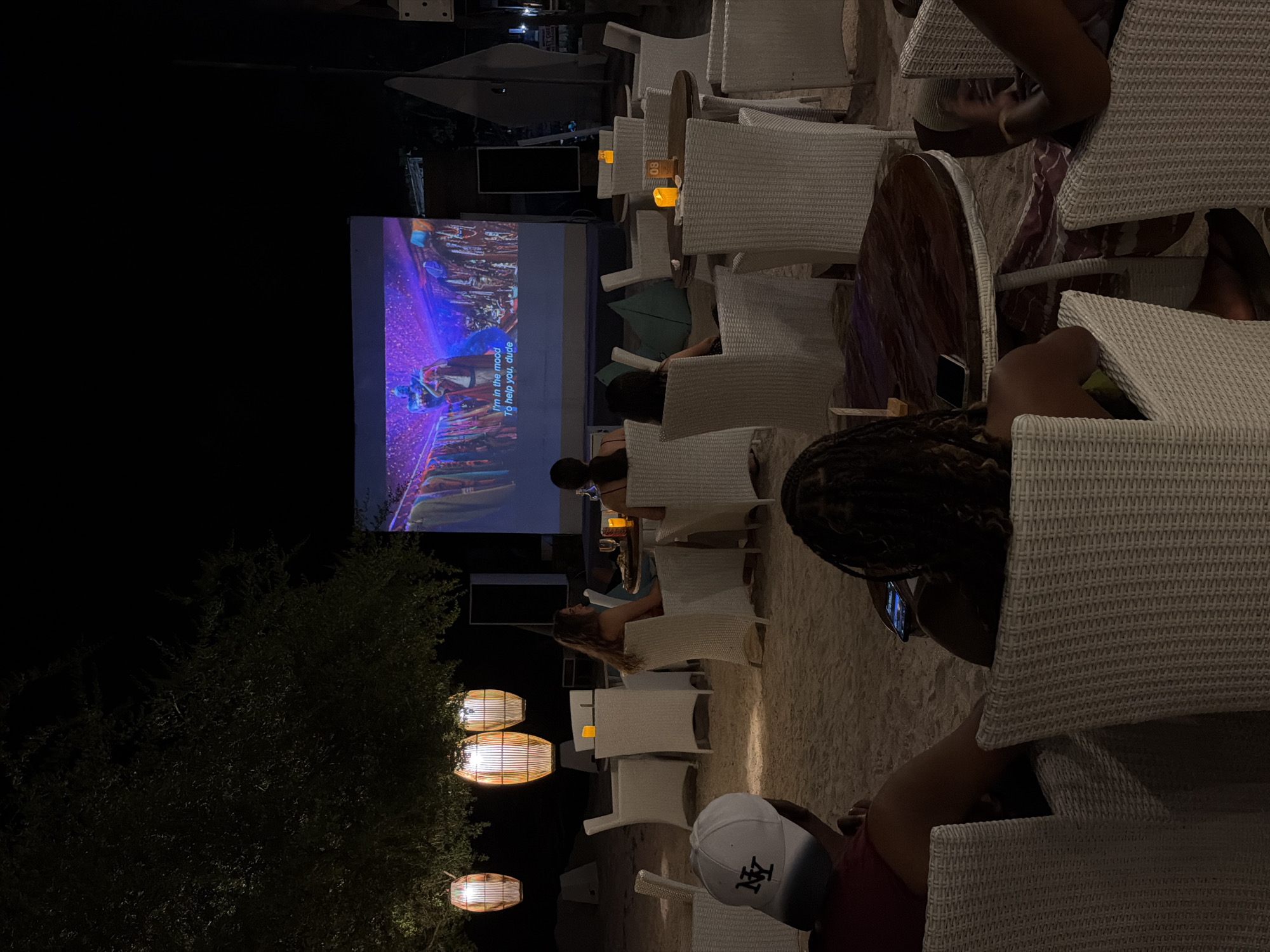 An outdoor cinema is set up on a sandy beach at night, with a large screen projecting a movie. Several white wicker chairs and tables are arranged on the sand, with some occupied by patrons. Three decorative lanterns hang from a tree, providing ambient light. A few tables have candles lit. The overall scene suggests a relaxed evening entertainment setting. The location is unknown but appears to be a coastal area in Indonesia.