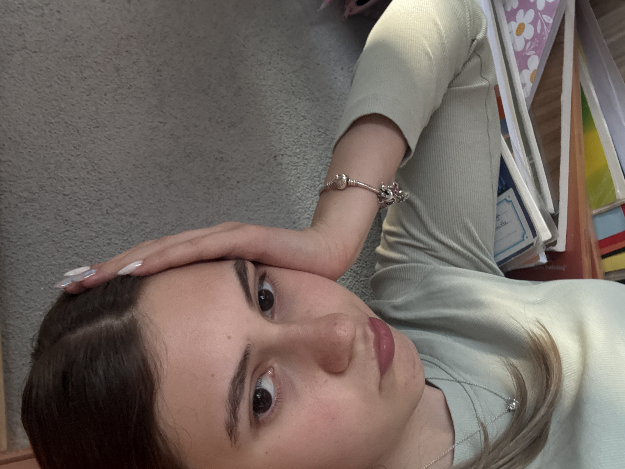 A close-up, high-angle shot depicts a young woman with fair skin and brown hair. She is wearing a light green, long-sleeved top. Her left hand rests on her forehead. A silver charm bracelet adorns her left wrist. A silver necklace is visible around her neck. The background includes a gray surface, possibly a carpeted floor or wall, and stacks of books and papers. There is no verifiable information suggesting the image was taken in Panagyurishte, Bulgaria, and the image provides no details or landmarks for location verification.