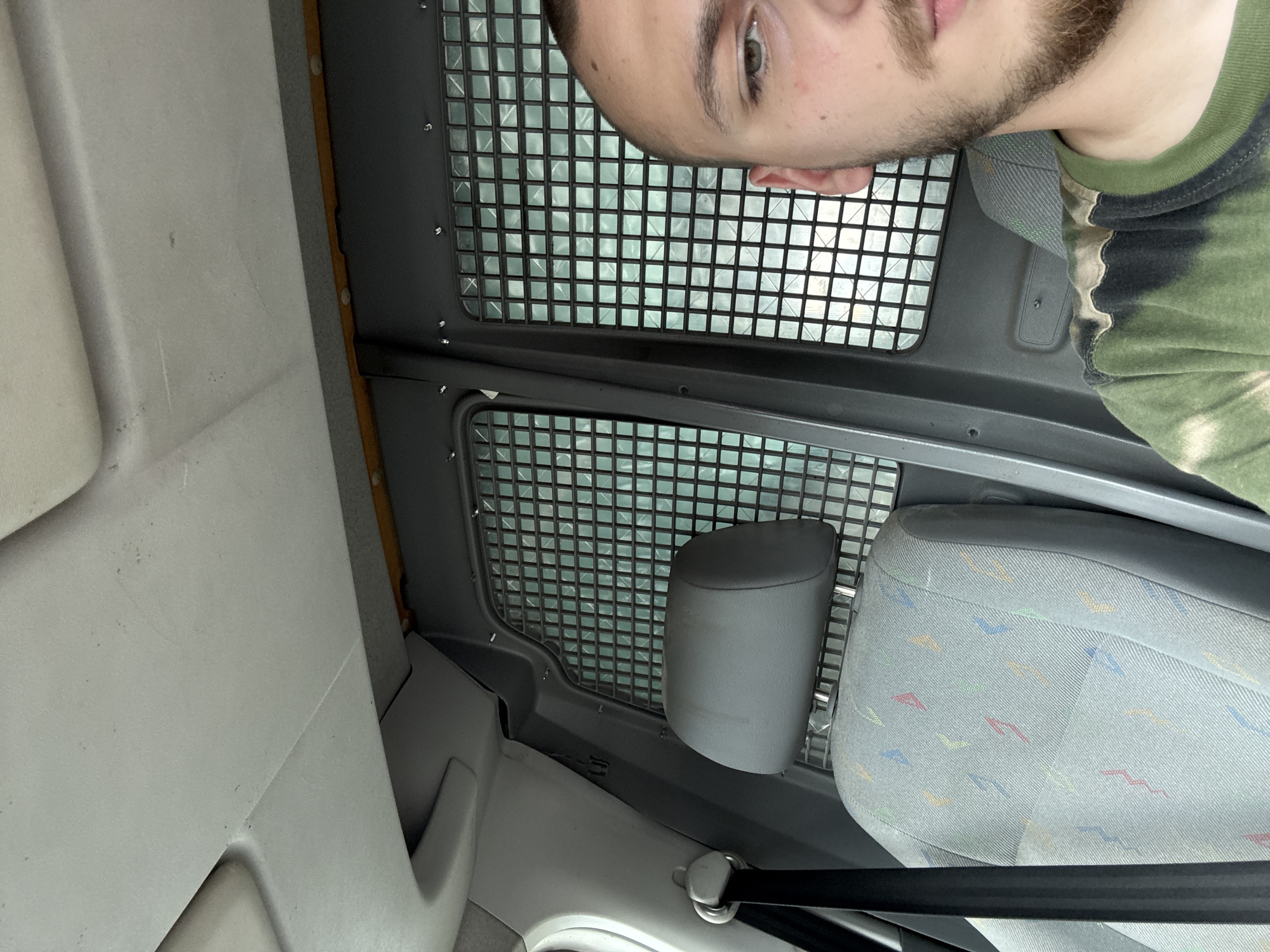 The image shows the interior of a vehicle, captured from a low angle, with a man's head and upper body positioned upside down in the upper right quadrant of the frame. The man has short dark hair and a beard, with his right eye looking towards the left. He is wearing a camouflage-patterned t-shirt in shades of green, black, and light tones.
A prominent dark grey metal partition with a square grid pattern spans across the central part of the image, suggesting the interior of a commercial van or similar utility vehicle, separating the front cabin from a cargo area. Bright light visible through the grid implies a view to the outside during daytime.
On the lower right, a grey fabric vehicle seat is visible, characterized by a colorful, abstract pattern of small dashes in blue, green, orange, and red. A black seatbelt with a metallic buckle is seen at the very bottom. Other interior components include plain grey plastic panels on the left side of the frame and a dark grey headrest or seat element partially visible through the metal grid. No text is visible in the image.