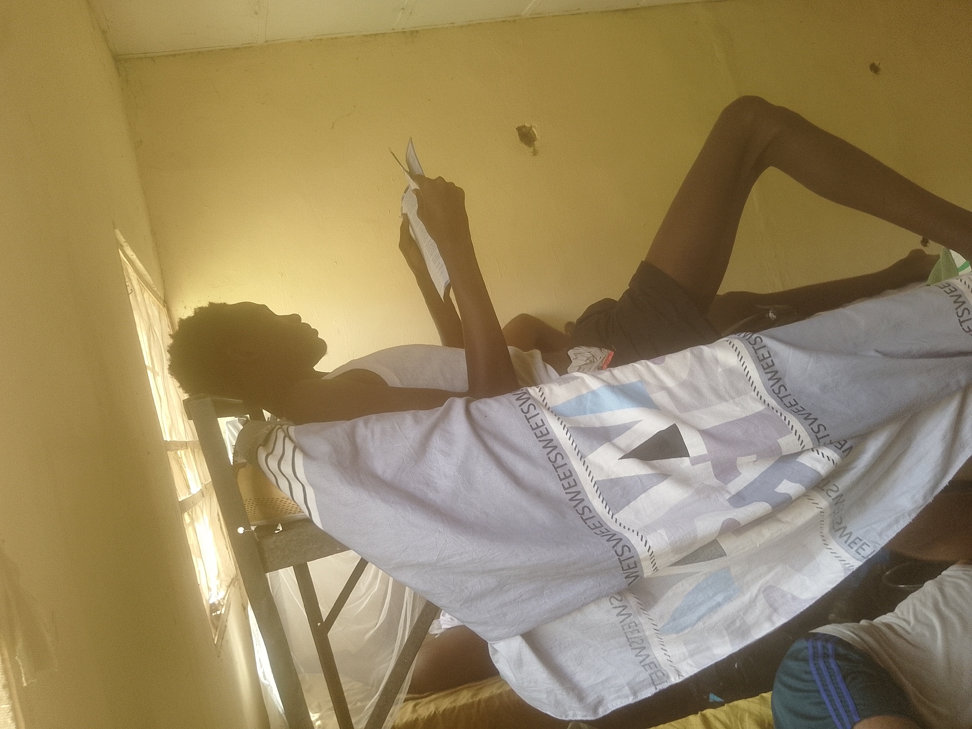 A person is lying on a bed, reading a book. They are positioned on their back, with their legs bent. The bed appears to be a bunk bed, as a metal frame is visible. The room is dimly lit, suggesting it could be evening or a room with limited natural light. The walls are a pale yellow. A window with a curtain is visible on the left side of the image, with sunlight streaming through, indicating it is daytime. The person is holding a book open with both hands and appears to be concentrating on reading. A light-colored blanket with a pattern of geometric shapes and text is spread over the bed. The visible text on the blanket reads "SWEETSWEETSW". The overall scene suggests a moment of quiet relaxation or study in a dormitory-like setting.