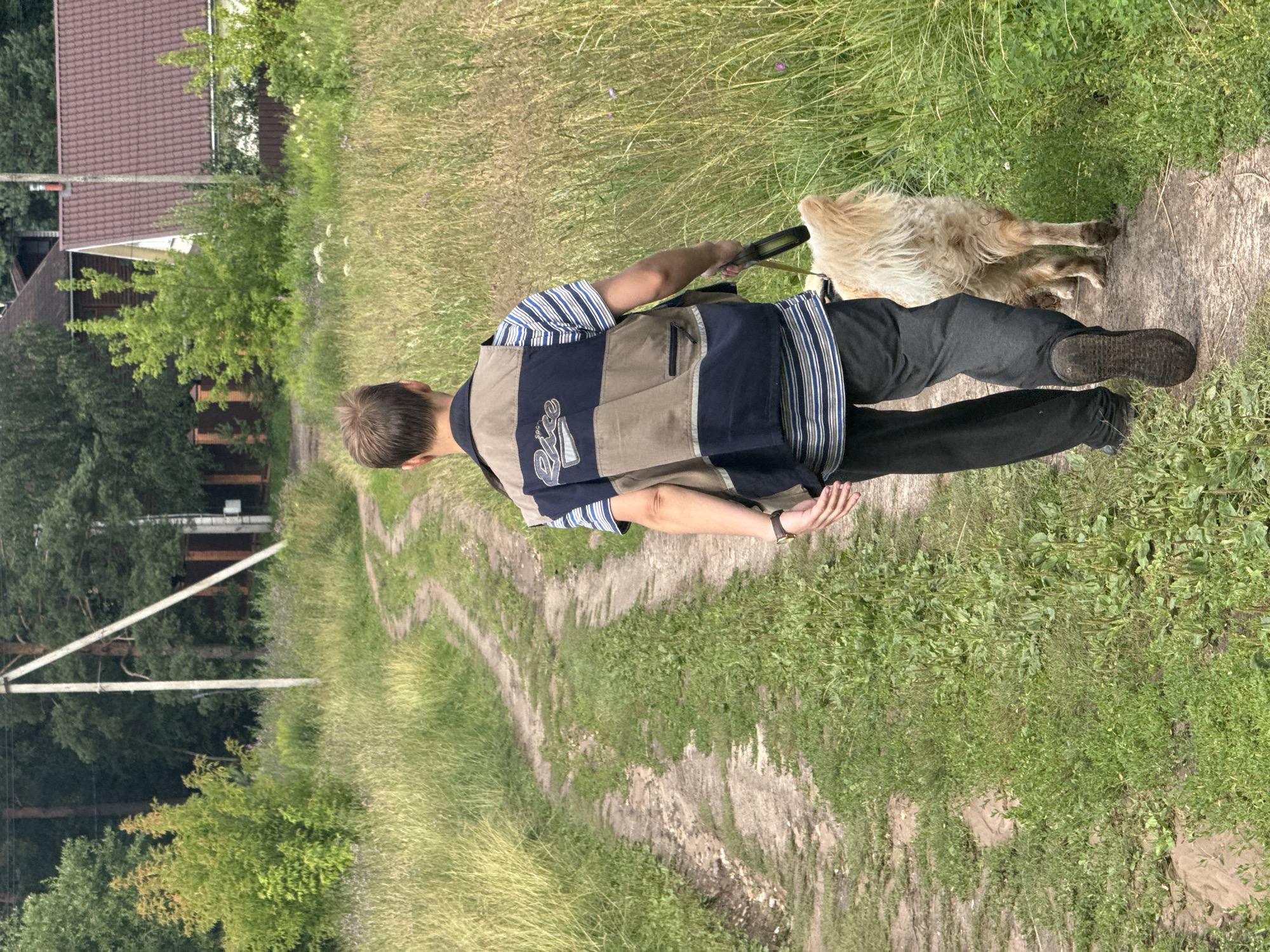 A person, viewed from behind, walks a shaggy, light-colored dog on a dirt path in a grassy, semi-rural area. The person is wearing a blue and white striped shirt under a tan and navy vest, and dark pants. They are holding a leash attached to the dog. The path winds through tall dry grass and appears to be a rural road. In the background, there are trees, houses with dark roofs, and a utility pole with wires. The setting suggests a location in the outskirts of Dmitrov, Russia, characterized by a blend of natural landscape and residential structures.