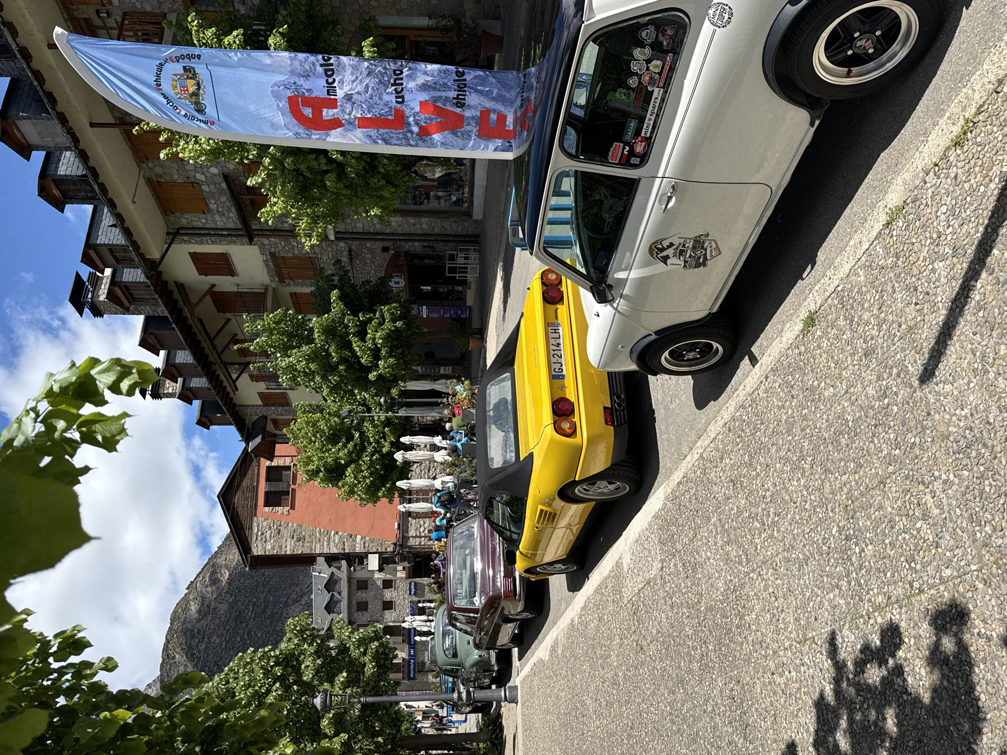 The image depicts a street scene in Benasque, Spain, with a row of parked vintage cars. The foreground features a white classic Mini Cooper with various stickers on its windows and body. Next to it is a yellow convertible, followed by a maroon car and others further down the street. A banner with the text "Amicale Véhicules Epoque" and a mountain range graphic is visible near the parked vehicles.
The buildings along the street are primarily constructed of stone and wood, exhibiting traditional architectural elements. Some buildings have balconies and shuttered windows. In the background, there is a restaurant with outdoor seating, where people are seated. The street is paved with cobblestones, and there are trees lining the street, partially obscuring the view. In the distance, mountains can be seen. The sky is blue with some clouds.