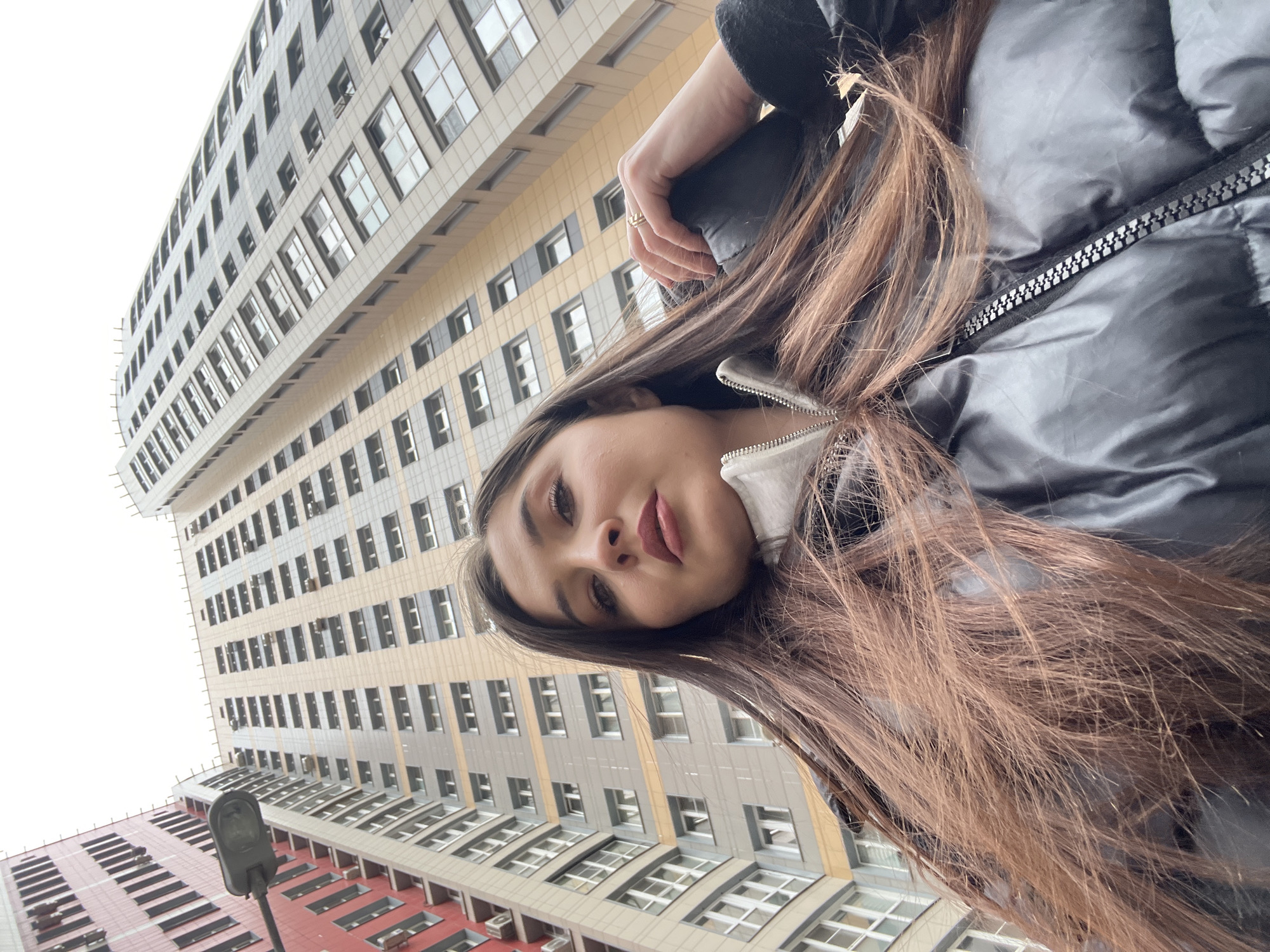 The image, captured from a low-angle perspective, features a young woman taking a selfie against a backdrop of tall, modern urban buildings. The woman is positioned in the foreground, her face looking directly into the camera with a neutral expression. She has long, dark brown hair that cascades across her shoulders and chest. She is wearing a black puffy jacket with a prominent silver zipper, and a white or light-colored collar is visible underneath. Part of her hand, adorned with a ring on her ring finger, is also visible.
The background is dominated by several multi-story buildings, characteristic of modern apartment or office blocks, extending high into a bright white sky. The buildings feature numerous rectangular windows and facades composed of light gray, beige, and a distinct reddish-brown section towards the bottom. The perspective emphasizes the height and scale of the structures, suggesting a bustling urban environment. A black streetlight fixture is partially visible in the lower left corner. The bright, possibly overcast sky indicates it is daytime. The architectural style and dense development are consistent with modern areas in a city like Khimki, Russia. There is no visible text in the image.