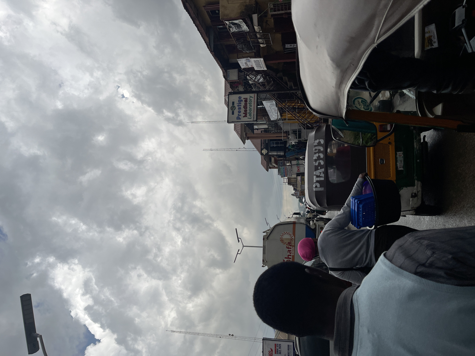 The image captures a busy street scene in Jos, Nigeria, under a cloudy sky. In the foreground, a person's head and shoulders are visible from behind, facing away from the camera, wearing a light blue vest over a plaid shirt. To their right, another person wearing a pink headscarf and a grey sweater is walking, carrying a large black basket filled with blue plastic containers. They are walking alongside a yellow and green auto-rickshaw (Keke Napep).
The street is congested with traffic, including the auto-rickshaw, a white tanker truck with "Shafa ENERGY" printed on its side, and other vehicles partially visible. Buildings line the right side of the street. One prominent building features a sign for "Prestige Medical Laboratory," with other smaller signs for "LUBES" and various businesses. The street appears to be wet, suggesting recent rain. Tall communication towers are visible in the background, indicating the urban setting. The overall impression is one of a bustling marketplace or transit hub in Jos.