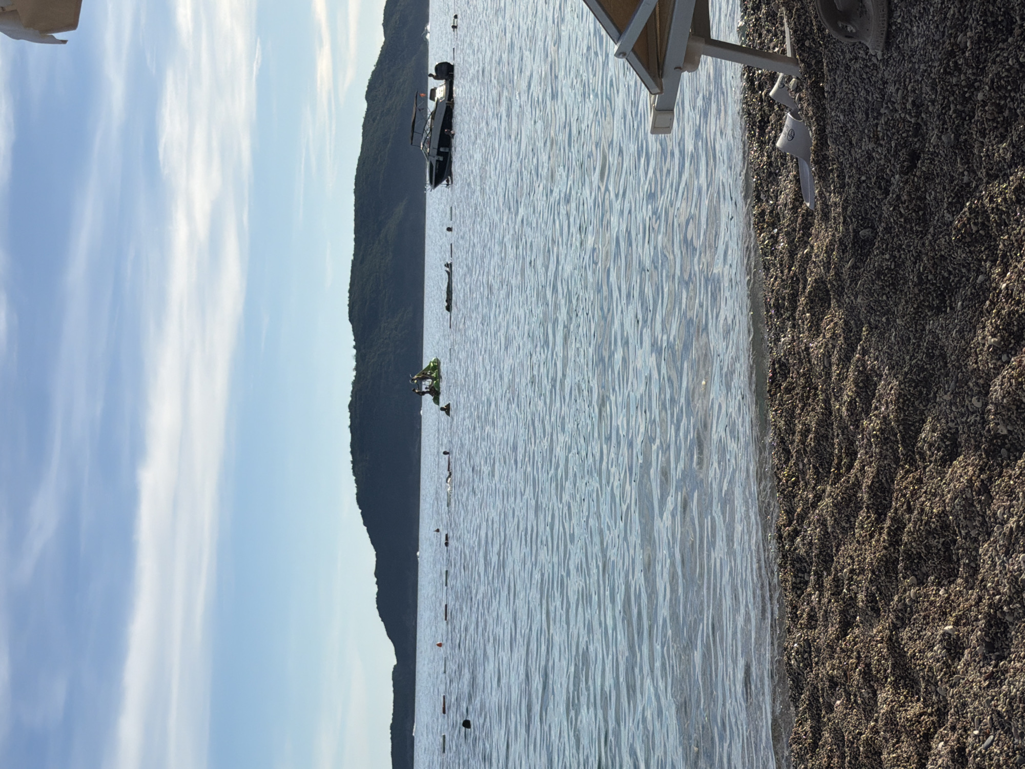 The image captures a serene coastal scene in Kamenovo, Montenegro. In the foreground, a pebbled beach stretches towards the water. A white beach chair is partially visible on the right, with a pair of white flip-flops resting in the sand near its base. The calm, blue sea fills the middle ground, with gentle waves lapping at the shore. Several buoys are visible, marking a swimming area. In the water, individuals can be seen swimming and engaging in water activities. A white boat with a dark canopy is anchored, and a jet ski or similar watercraft is present further out. In the background, a lush green, treed hill forms a natural backdrop against the clear blue sky, which is adorned with wispy white clouds. The overall atmosphere is one of peaceful relaxation and outdoor recreation.