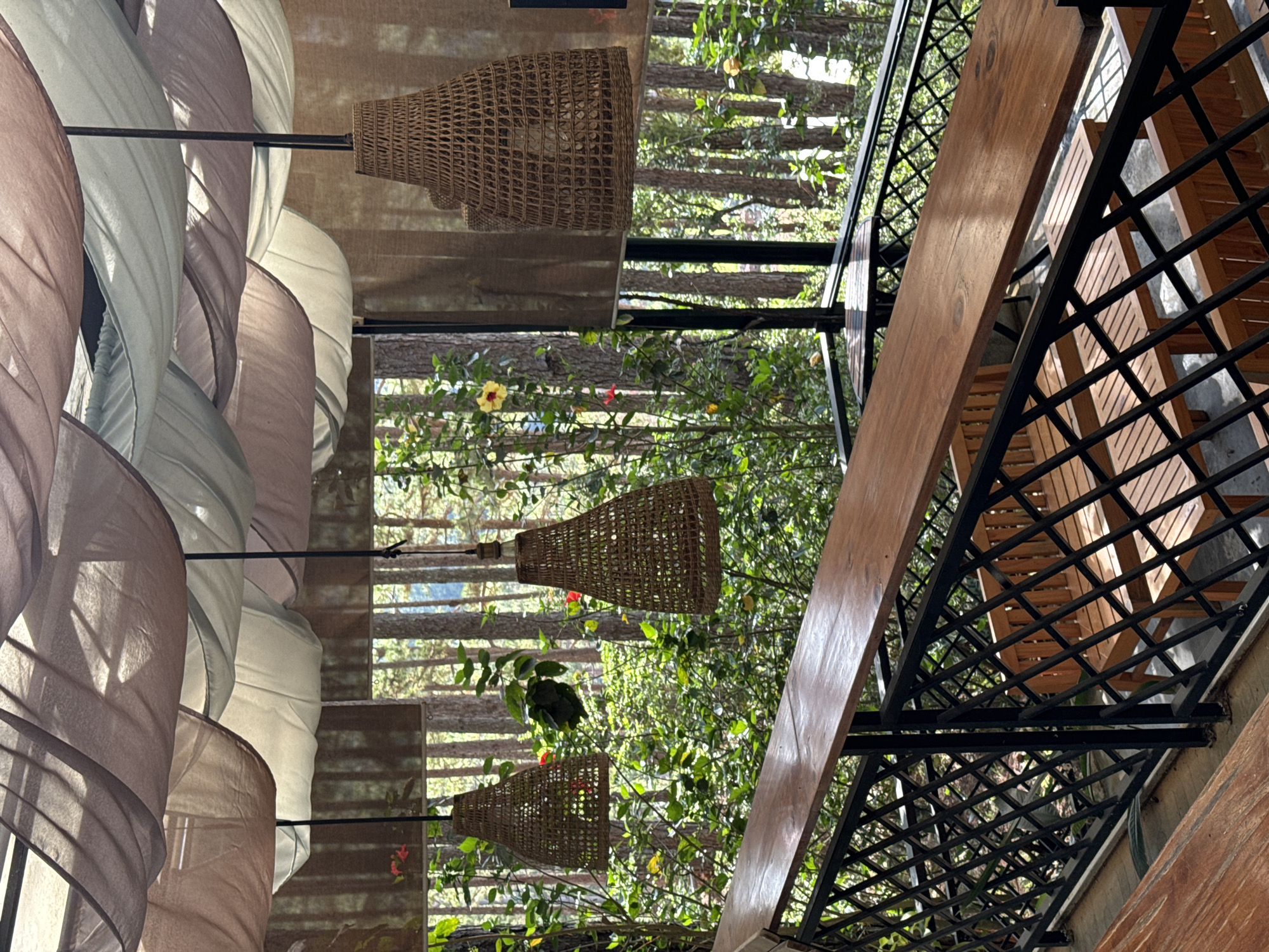 The image shows an open-air structure, possibly a restaurant or cafe, in Da Lat, Vietnam. The view features suspended woven light fixtures, sheer fabric panels hung overhead, and a section of outdoor seating with wooden benches and black metal railings. Through the open walls, there is dense foliage, suggesting a location surrounded by trees.