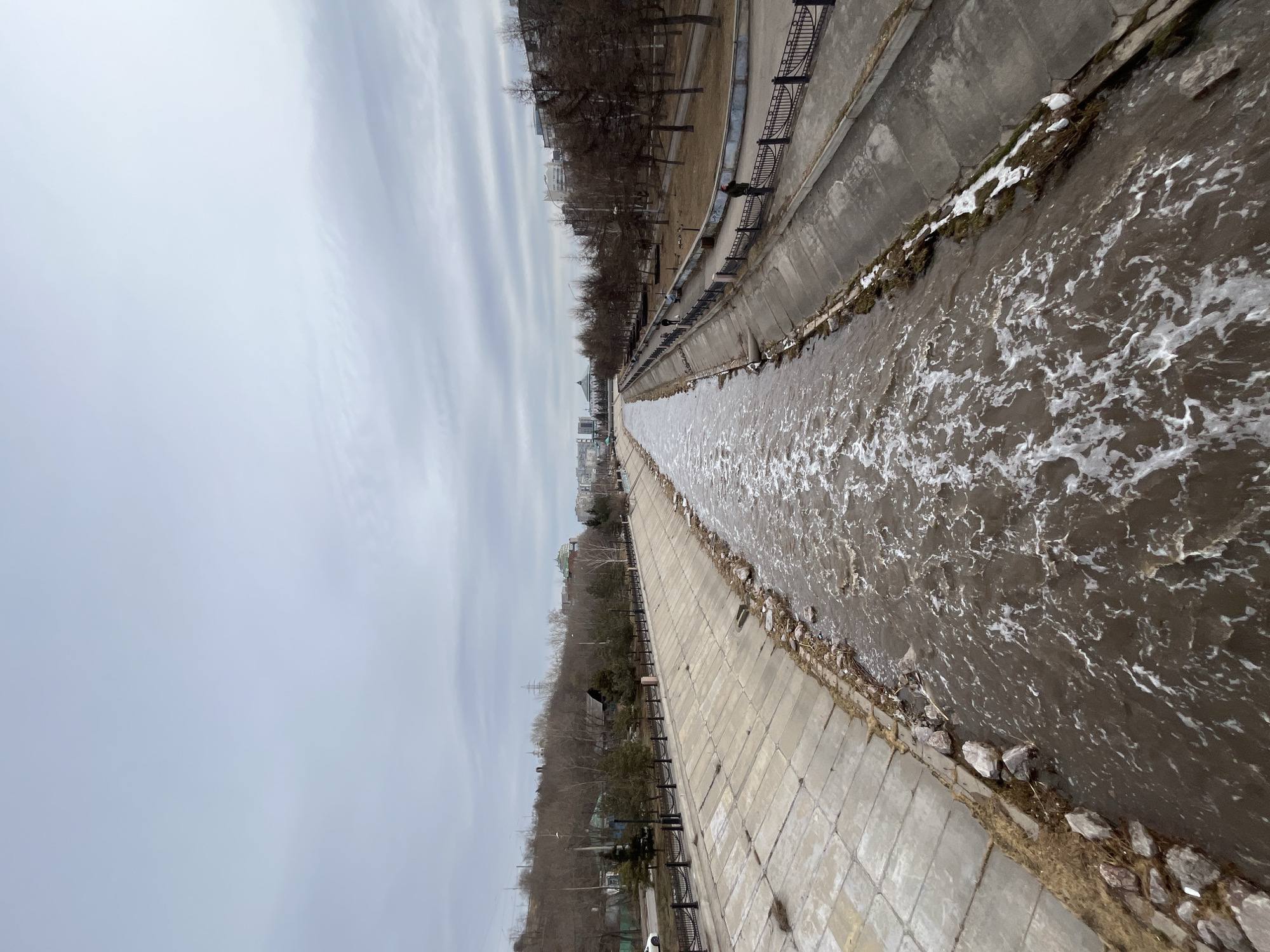 The image displays a channelized section of the Kacha River in Krasnoyarsk, Russia, viewed longitudinally from an elevated position looking downstream. The environment indicates late winter or early spring conditions.
The Kacha River occupies the central portion of the frame, characterized by rapidly flowing, turbulent, brown water with extensive white foam and aerated patches. Its banks are uniformly constructed from large, sloped concrete panels. Minor deposits of dried vegetation and scattered rocks are visible along the water's edge on both sides, with small patches of snow present on the right bank.
The left bank consists of the concrete channel wall, an elevated paved walkway, and a continuous black metal fence with decorative finials. Beyond this fence lies a narrow strip of dry, brown ground, followed by a road on which a white sedan is partially visible. Further uphill, a steep embankment rises, covered primarily with bare deciduous trees, interspersed with some evergreen conifers and utility poles carrying power lines. Distant urban buildings are visible atop this incline.
The right bank parallels the left, featuring the concrete channel wall, an adjacent paved walkway, and a similar black metal fence. Beyond the fence, a park area extends, dominated by dry, brown grass and numerous bare deciduous trees. Streetlights are regularly spaced within this park. Three individuals are present on the right bank: one person in a dark coat stands near the fence in the mid-ground, another figure walks along the paved path further into the distance, and a third individual is partially visible deeper within the park area. In the distant background on the right, various urban buildings are discernible, including a prominent structure with a large, light-colored domed roof, which is the Krasnoyarsk State Opera and Ballet Theatre.
The sky is overcast and uniformly light grey, showing subtle banding in its cloud cover.