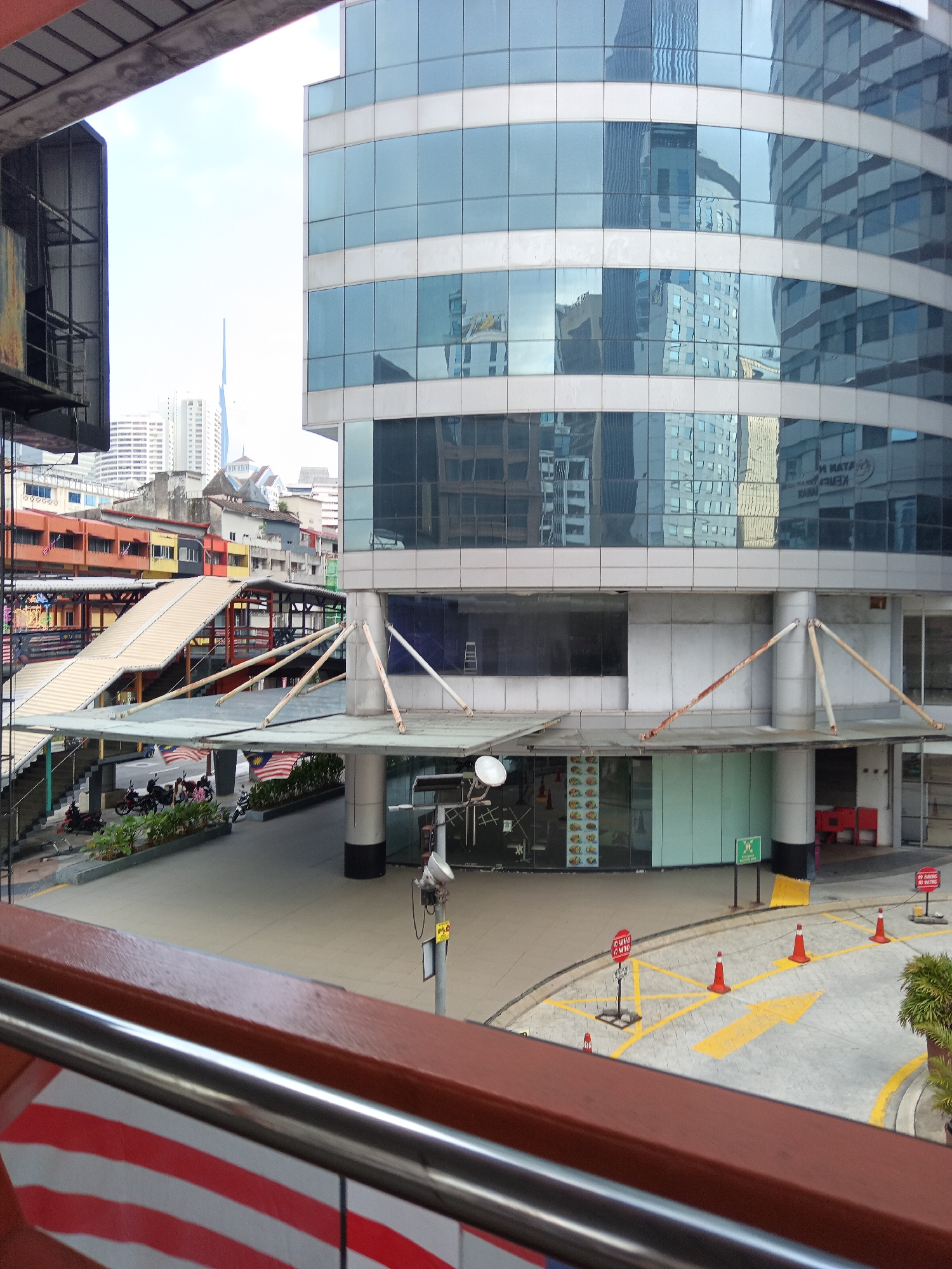 The image depicts an elevated view of a street and pedestrian walkway in Kuala Lumpur, Malaysia, likely in the Bukit Bintang area, given the architectural style and presence of Malaysian flags. In the foreground, a reddish-brown railing with a silver handrail runs horizontally across the bottom of the frame, adorned with red and white striped patterns.
Beyond the railing, a multi-level pedestrian walkway with tan-colored steps and yellow handrails connects to a modern building with a glass facade. This building features multiple stories of reflective blue glass windows interspersed with white architectural elements. The glass reflects other buildings and the sky. Beneath the walkway, a street level area is visible with parked motorcycles, potted plants, and a paved surface marked with yellow lines and arrows. Orange traffic cones are strategically placed on the paved area, and red signs indicating "NO PARKING" are visible.
The background reveals a dense urban landscape with numerous high-rise buildings of various designs and colors. The sky is overcast, suggesting a cloudy day. The overall scene captures a bustling urban environment with a blend of modern architecture and older structures, indicative of a dynamic city like Kuala Lumpur. There are no discernible people in the immediate foreground or mid-ground, but the presence of parked vehicles implies activity in the area.