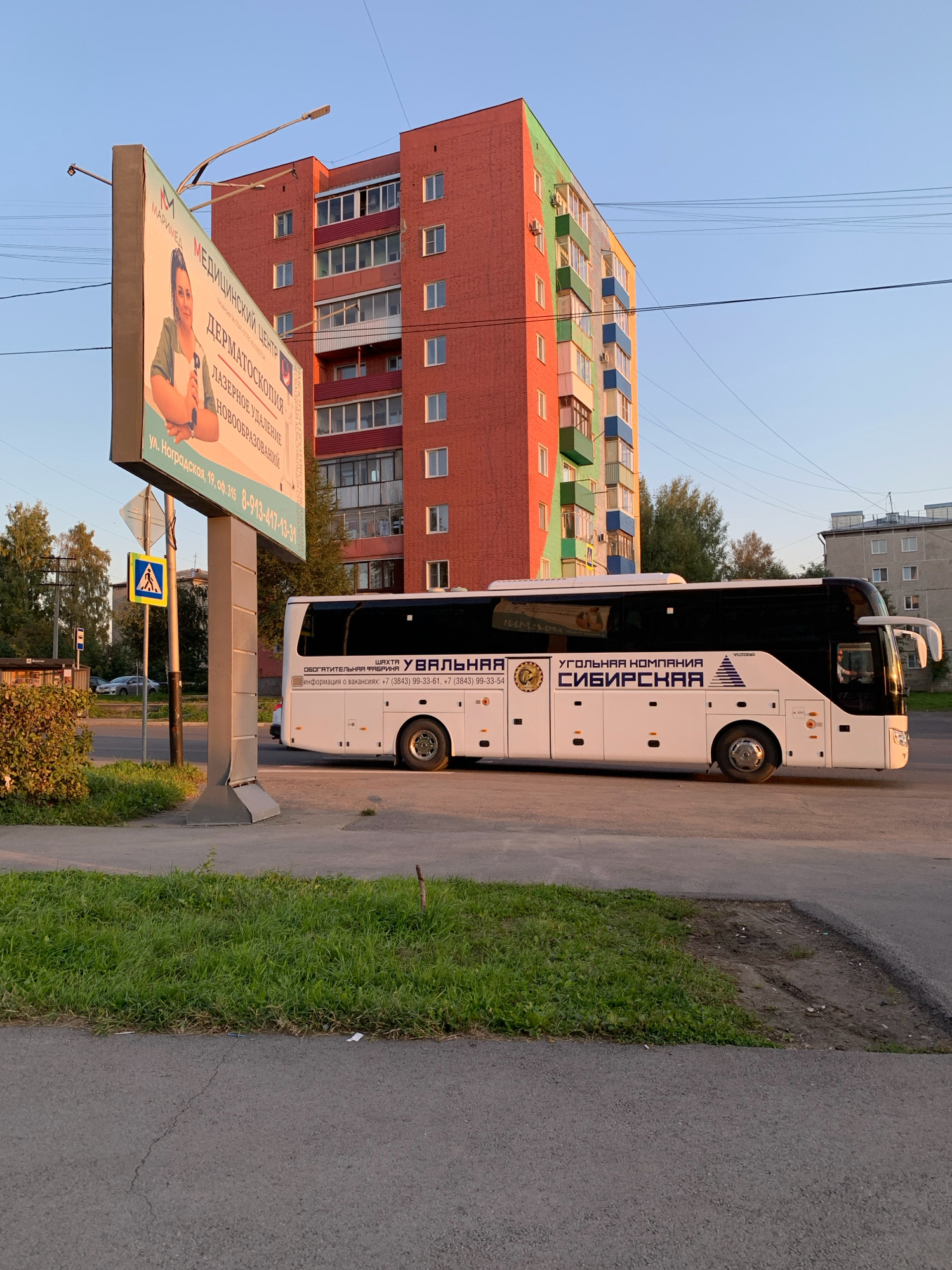 The image shows a white bus with "УГОЛЬНАЯ КОМПАНИЯ СИБИРСКАЯ" (COAL COMPANY SIBIRSKAYA) written on its side. The bus is parked on a street in front of a multi-story brick building in Prokopyevsk, Russia. The building has a colorful facade with sections of red, green, and blue. A billboard on the left advertises a medical center. The sky is clear and blue, and the sun is setting, casting a warm light on the scene. The area is urban, with trees and other buildings visible in the background.