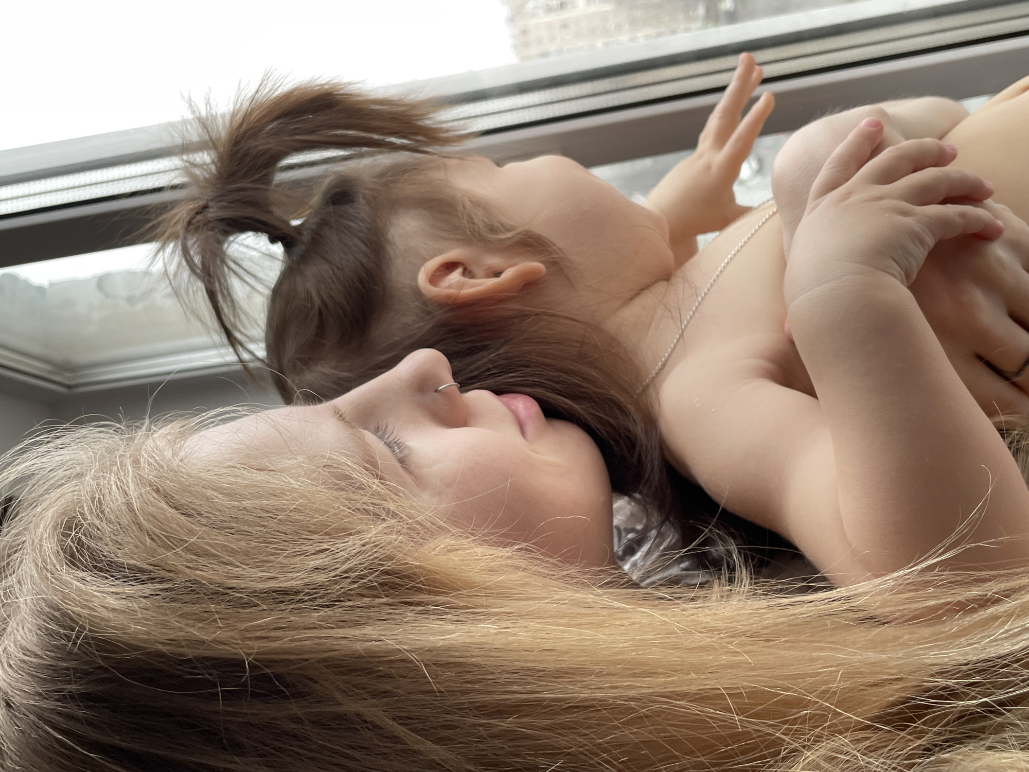 A woman with long blonde hair is lying down with a young child with brown hair pulled into a ponytail. They are both looking upwards towards a window, and the child's hands are raised. A city skyline is visible through the window. The exact location in Izhevsk, Russia cannot be determined from the image.