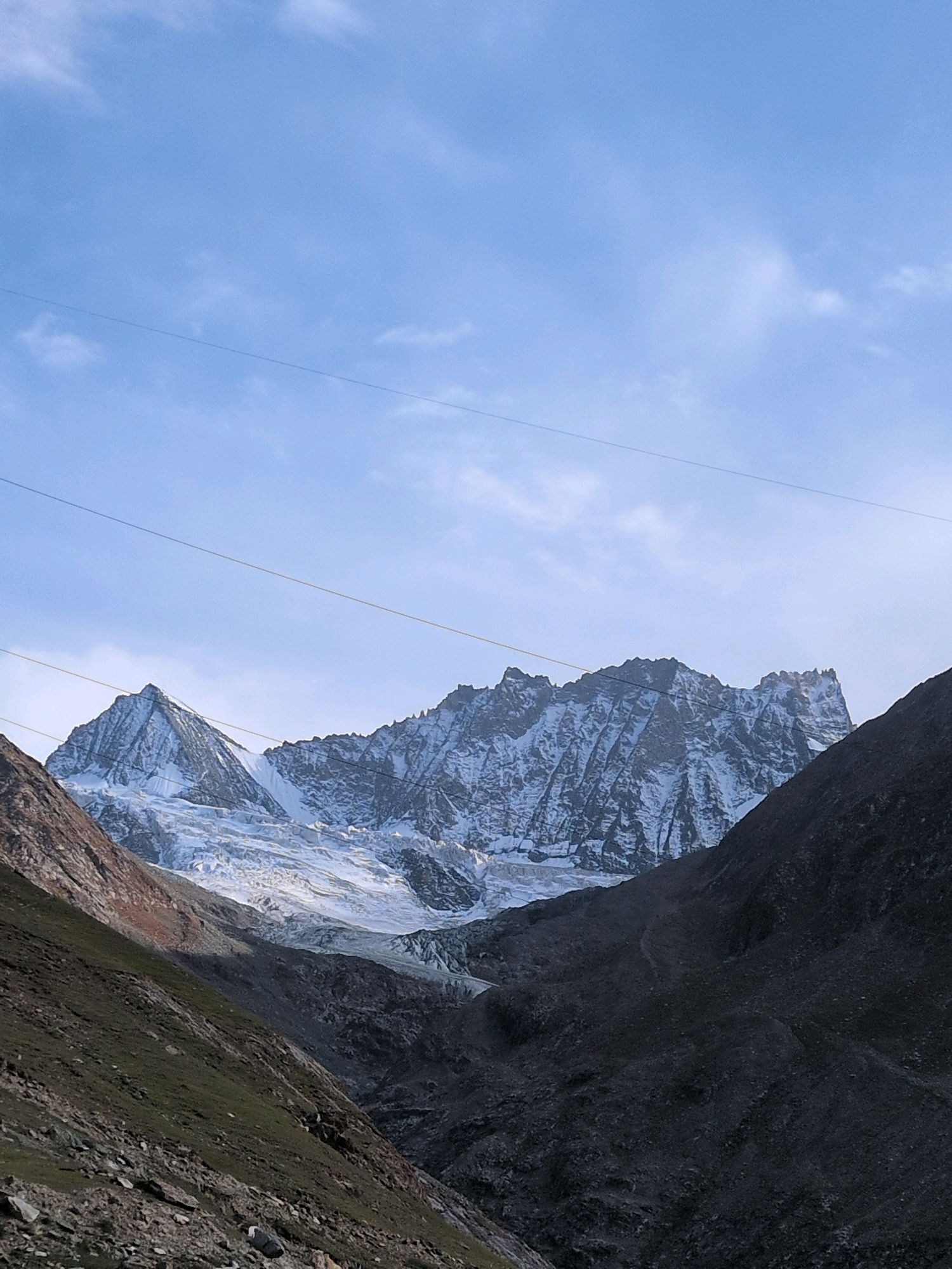 The image shows a majestic mountainous landscape in India. In the foreground, a steep, green-covered slope descends into a valley, with rocks scattered across the terrain. The middle ground is dominated by a massive snow-covered mountain range, its peaks jagged and sharp against the sky. A prominent glacier flows down the mountain, reflecting the light. The background features a clear blue sky with a few wispy clouds, and two power lines stretch across the upper portion of the frame. The scene appears to be captured during daylight, with the sun casting shadows that highlight the rugged textures of the mountains. No people or man-made structures, other than the power lines, are visible in the image.
