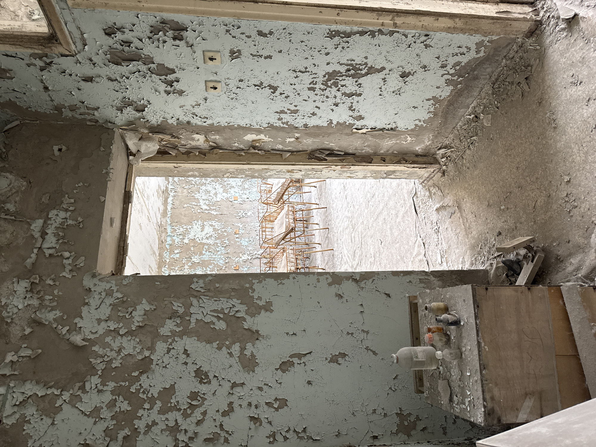 The image depicts the interior of an abandoned room in what is likely a building in Pripyat, Ukraine. The walls and ceiling are heavily damaged, with extensive peeling paint revealing underlying layers. A doorway leads to another room with similar conditions, where a rusted metal structure is visible. On a surface within the room, several glass bottles and containers are arranged. The room appears derelict, with accumulated dust and debris covering surfaces. There is no evidence of recent activity.