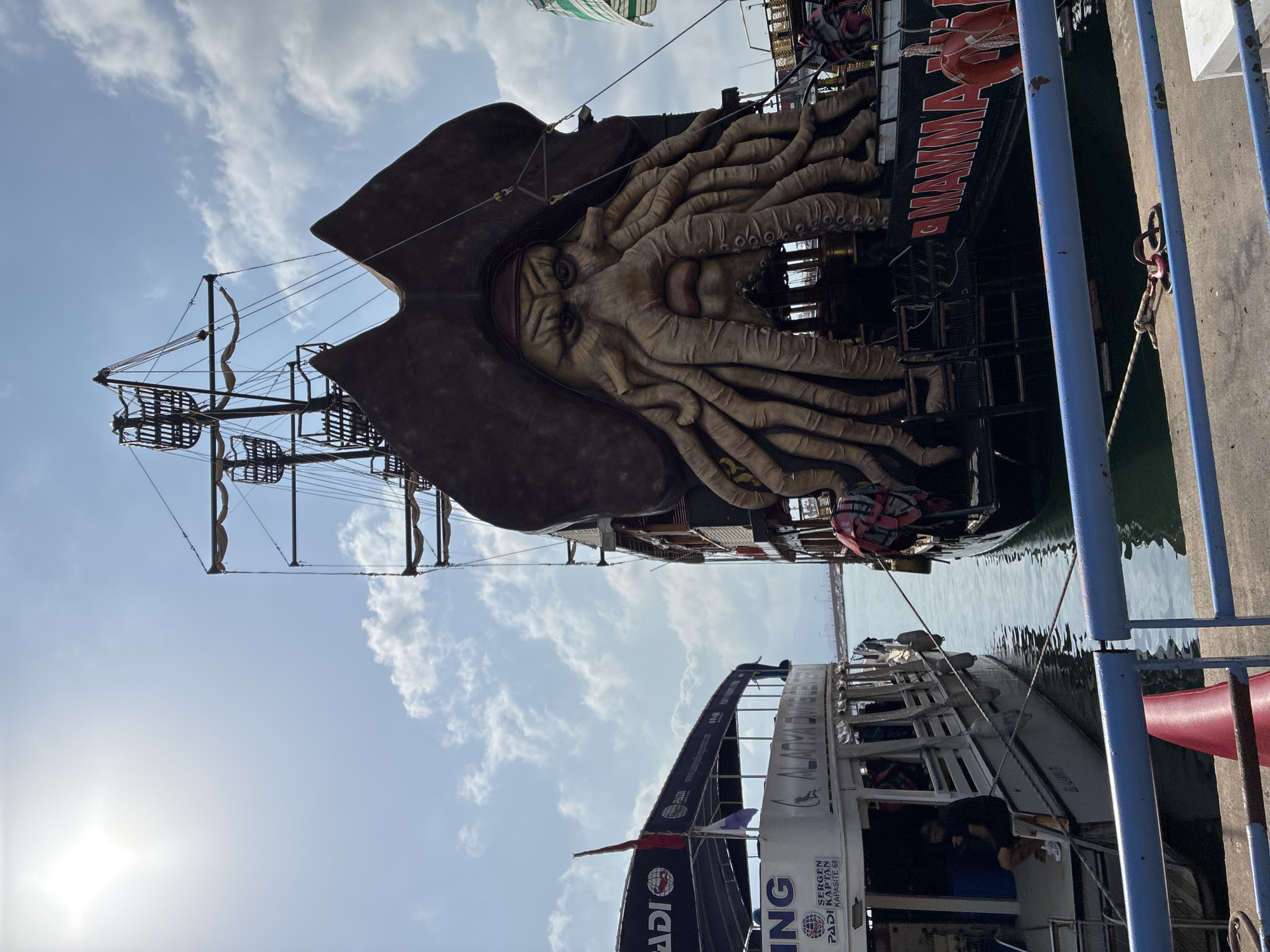 In Alanya, Turkey, two boats are docked side-by-side on a bright, sunny day with scattered clouds. The boat on the right is a pirate-themed vessel, featuring a large, elaborate mask of a sea creature with tentacles adorning its bow. The mask has a menacing expression with wide eyes and a prominent nose. The boat is dark-colored with "MAMMA MIA" written in large red letters on its side. A life preserver and a red life vest are visible. The boat on the left is a white passenger ferry with a covered seating area. It displays "PADI" and "ALANYA DIVE CENTER" on its exterior. A flag is visible flying from its mast. A few passengers are seated on the ferry. The boats are in the water, and a concrete dock with blue railings is in the foreground. The sun is shining brightly, creating a glare in the sky.