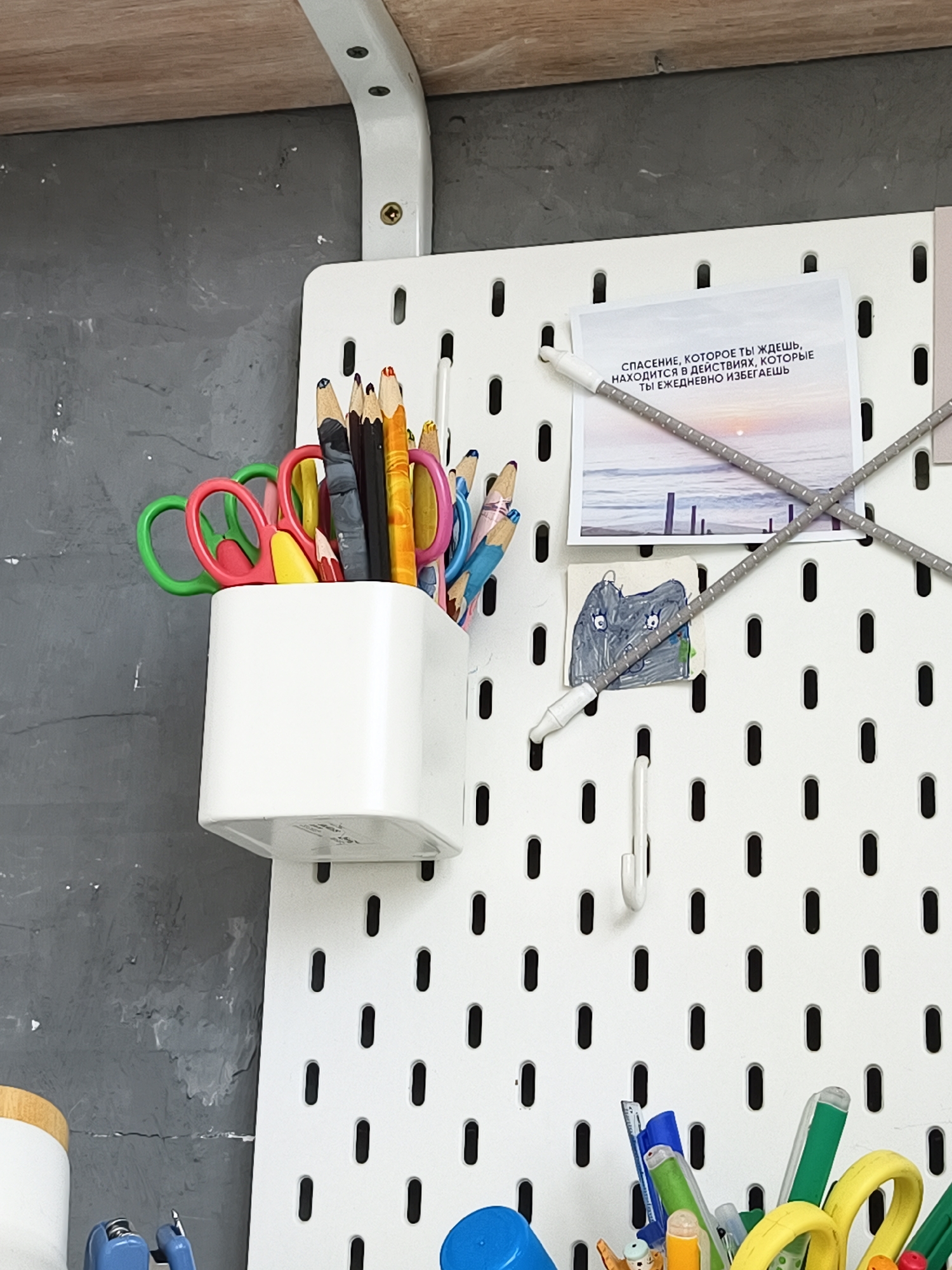 The image shows a close-up of a white pegboard wall mounted on a gray, textured wall. A wooden shelf is visible at the top of the frame. On the pegboard, a white plastic container holds various colored pencils and small scissors. To the right of the container, a postcard with text in Russian is attached to the pegboard with two grey elastic cords. Below the postcard is a small, child-like drawing of an elephant. A white hook is also attached to the pegboard. The lower portion of the image shows more writing utensils in a container, including pens and markers, and a blue object. The overall scene suggests an organized workspace or craft area. The location is stated to be in Volzhsk, Russia.