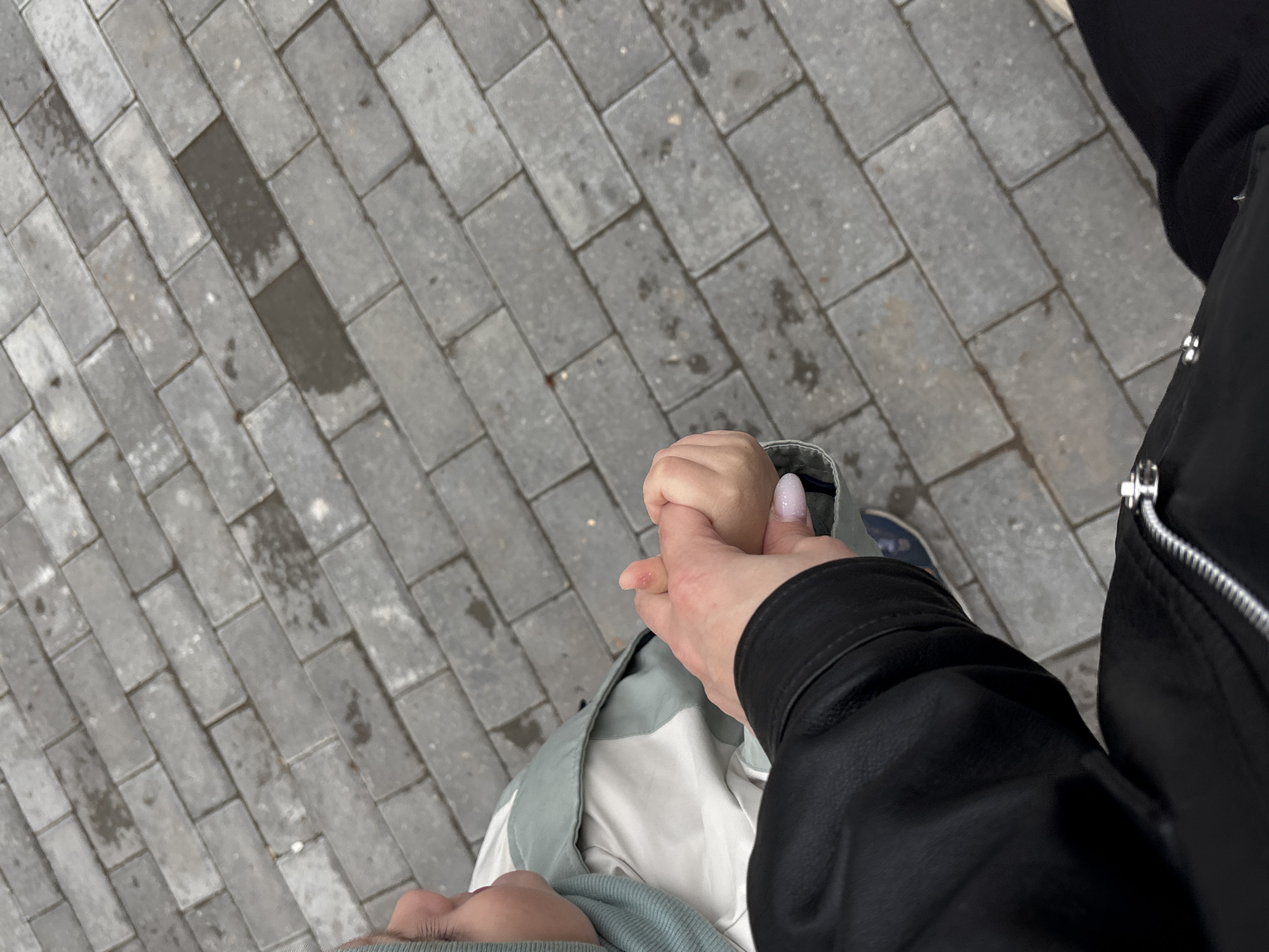 This image presents a downward, first-person perspective of two individuals holding hands while walking or standing on a paved surface. On the right, an arm clothed in a black jacket, likely leather, extends, with its hand gently holding the smaller hand of another person. This second individual, positioned on the left side of the frame, appears to be a child, wearing a light green and white jacket over a light-colored garment. Their head is tilted downwards, obscuring their face, and their fingernails are visible, painted a light, possibly glittery, color.
The ground is paved with grey, rectangular interlocking stones arranged in a diagonal pattern. Patches of the paving stones are visibly damp or wet, suggesting recent rain or humid conditions. In the background, a glimpse of a light blue or grey shoe is visible near the child's leg. The scene takes place outdoors, possibly on a sidewalk or path in Chaikovsky, Russia. The act of holding hands conveys a sense of connection, care, or companionship, likely an adult guiding a child. The lighting suggests daytime, possibly under an overcast sky. There is no visible text in the image.