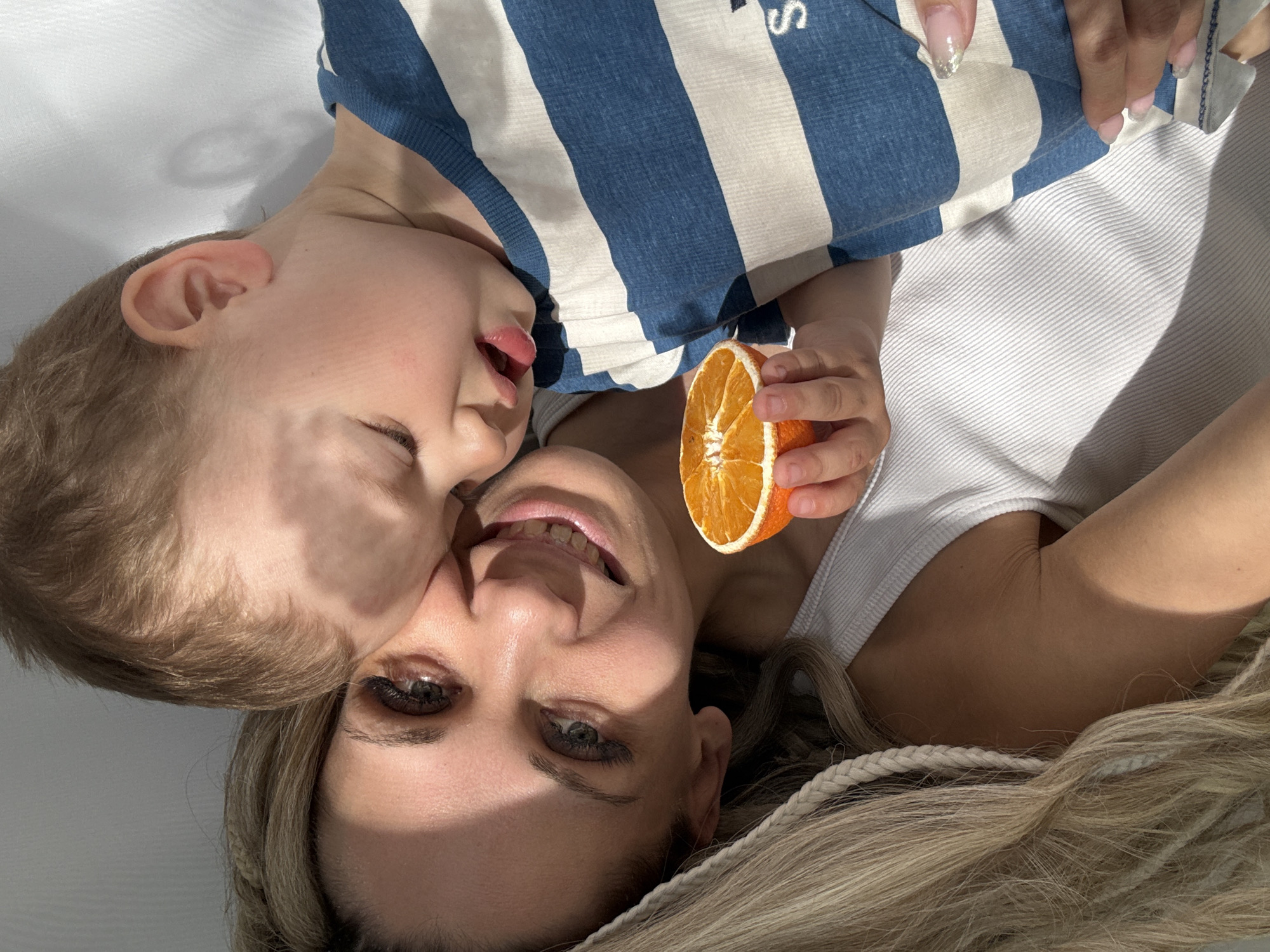 A woman with blonde hair and green eyes is lying on her back, smiling at the camera, with a young child lying on top of her. The child has blonde hair and is wearing a blue and white striped shirt. The woman is holding half of an orange. The background is a plain white surface. The location cannot be determined from this image.