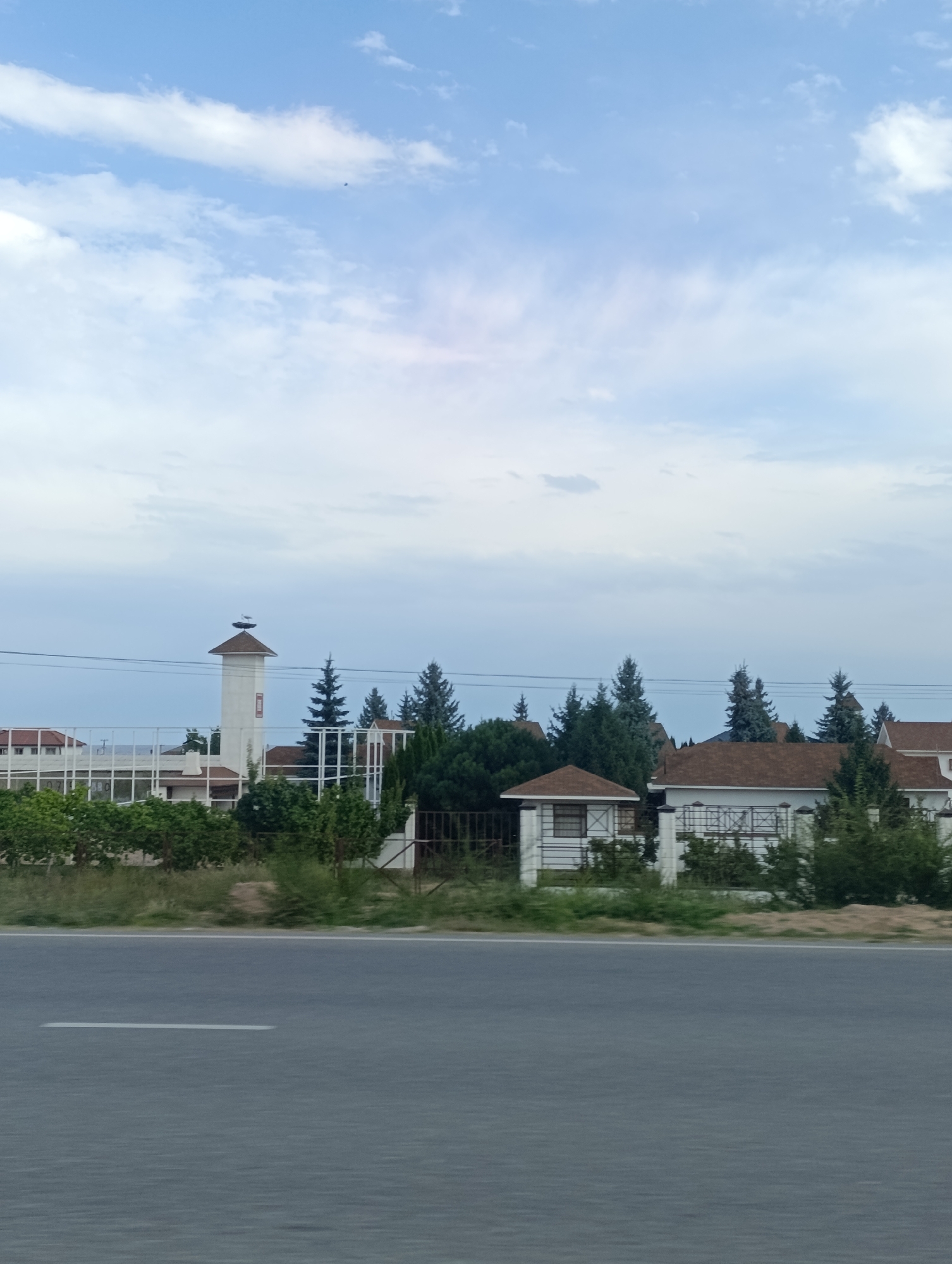 The image shows a view of buildings and trees on a cloudy day, likely in the city of Tamchy, Kyrgyzstan. In the foreground, there is a grey asphalt road with a white stripe. Behind the road, a white fence separates the viewer from a grassy area with various bushes and trees. To the left, there is a tall, white tower with a brown conical roof, on top of which two storks are visible. Further behind this tower, more buildings with white walls and brown roofs can be seen, along with a white fence and lush greenery. To the right of the tower, several coniferous trees stand tall amongst other residential buildings. The sky is filled with scattered white and grey clouds, with a clear blue sky peeking through in some areas. There are no people visible in the image.