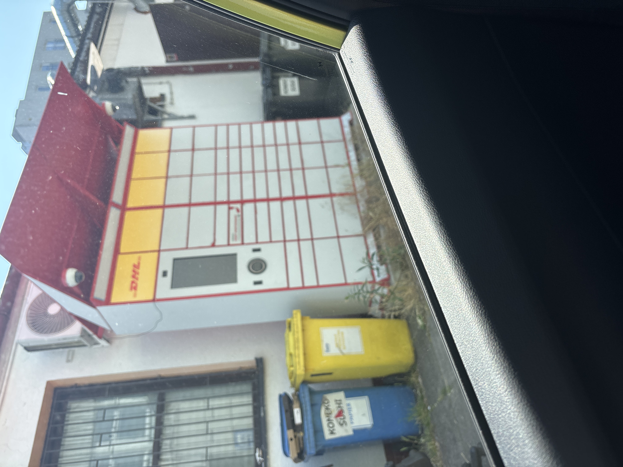 The image shows a DHL parcel locker station situated on a street in Krakow, Poland. The parcel locker is white with red and yellow accents and a red roof, indicating it is a DHL branded unit. It is positioned next to a building with a barred window and an air conditioning unit. In front of the parcel locker, there are two trash bins, one blue and one yellow, with visible labels. The scene appears to be outdoors on a sunny day, with the view framed from inside a vehicle, suggesting the photo was taken from a car. The environment shows some dry grass and vegetation around the bins and locker.